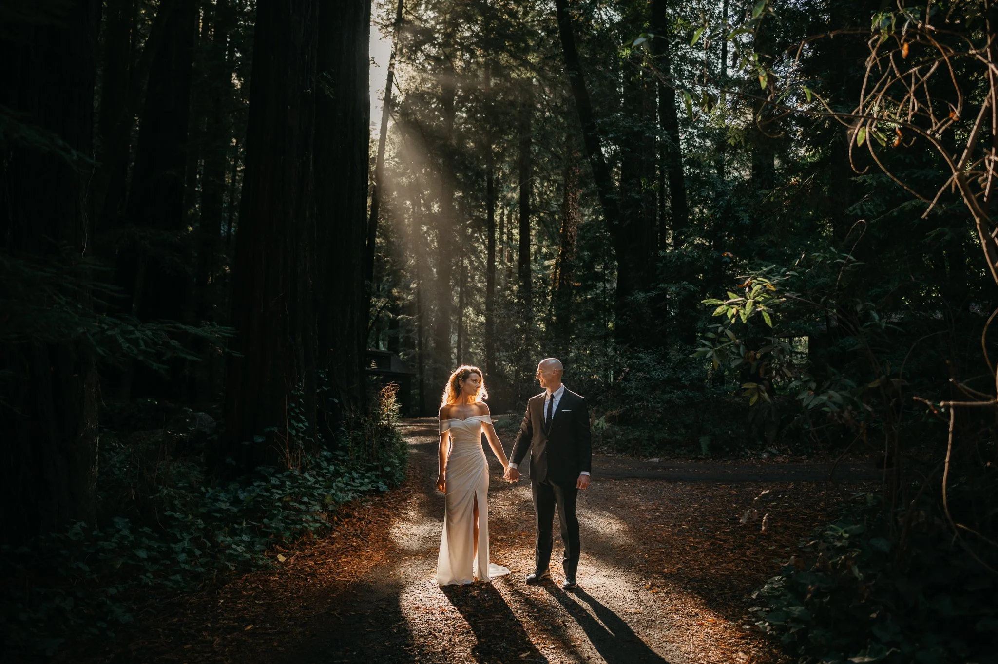 A bride and groom holding hands walking in a forest during sunset, with sunlight streaming through trees.