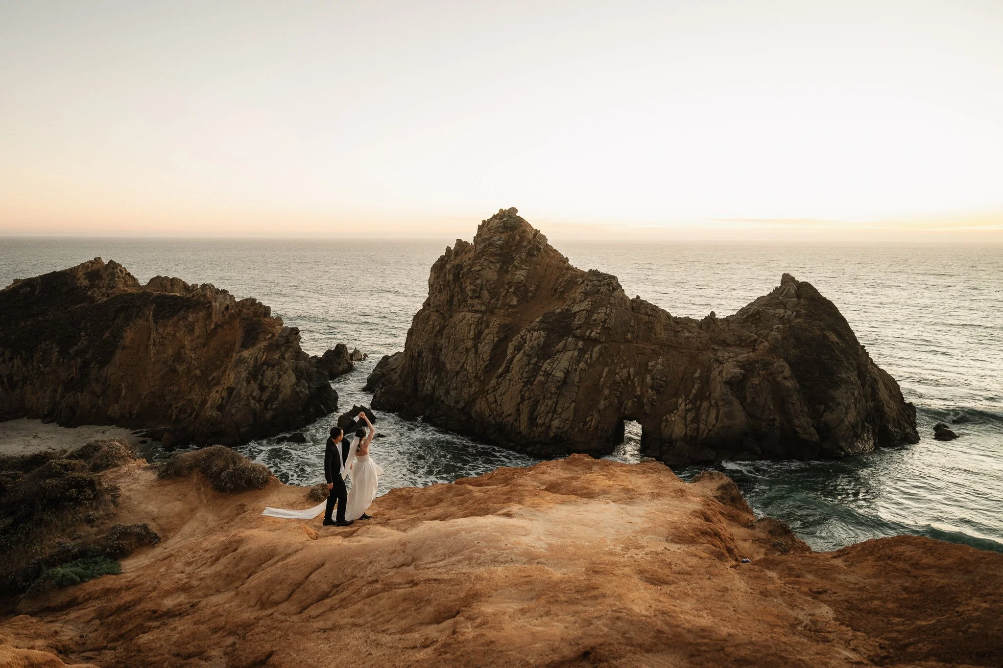 A bride and groom in wedding attire standing on a rocky cliff near the ocean at sunset, with large rocks in the background and the sea stretching to the horizon.
