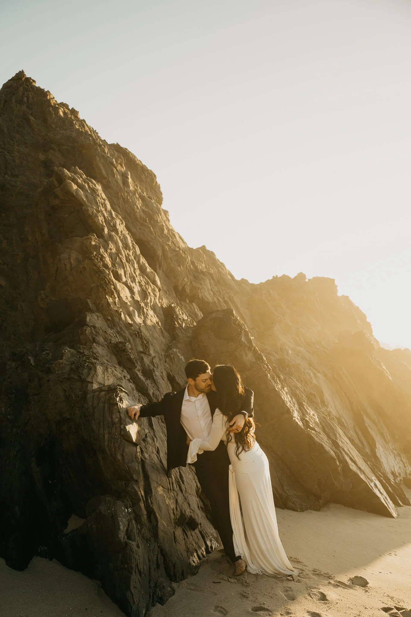 A couple dressed in formal attire sharing a kiss on the beach at sunset, with large rocks and a sandy shoreline in the background.
