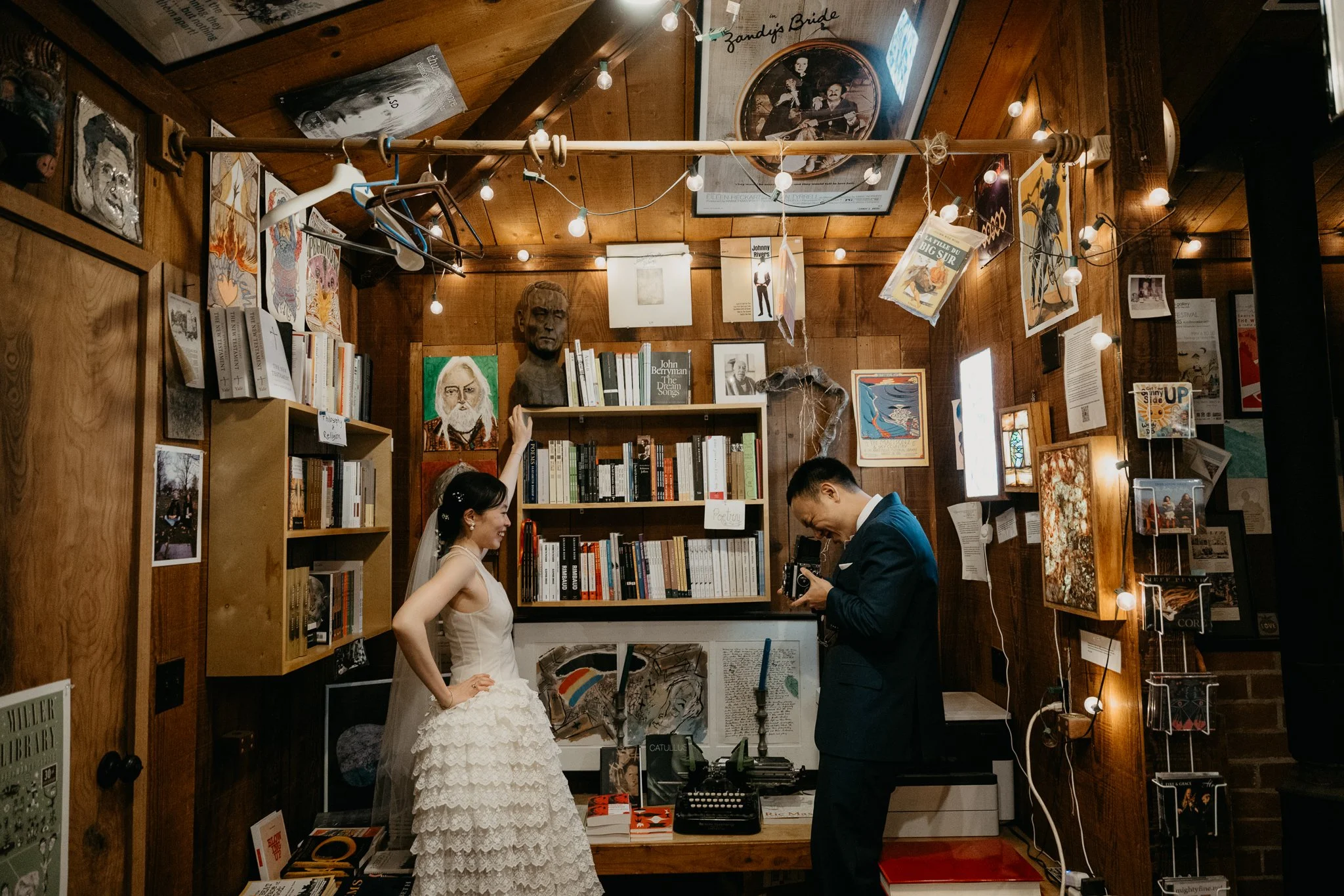 A bride in a wedding dress with lace ruffles and a veil, standing next to a groom in a suit, both inside a cozy, decorated room filled with books, artwork, and string lights. The bride is smiling and pointing at a bookshelf, while the groom is looking down at a camera.