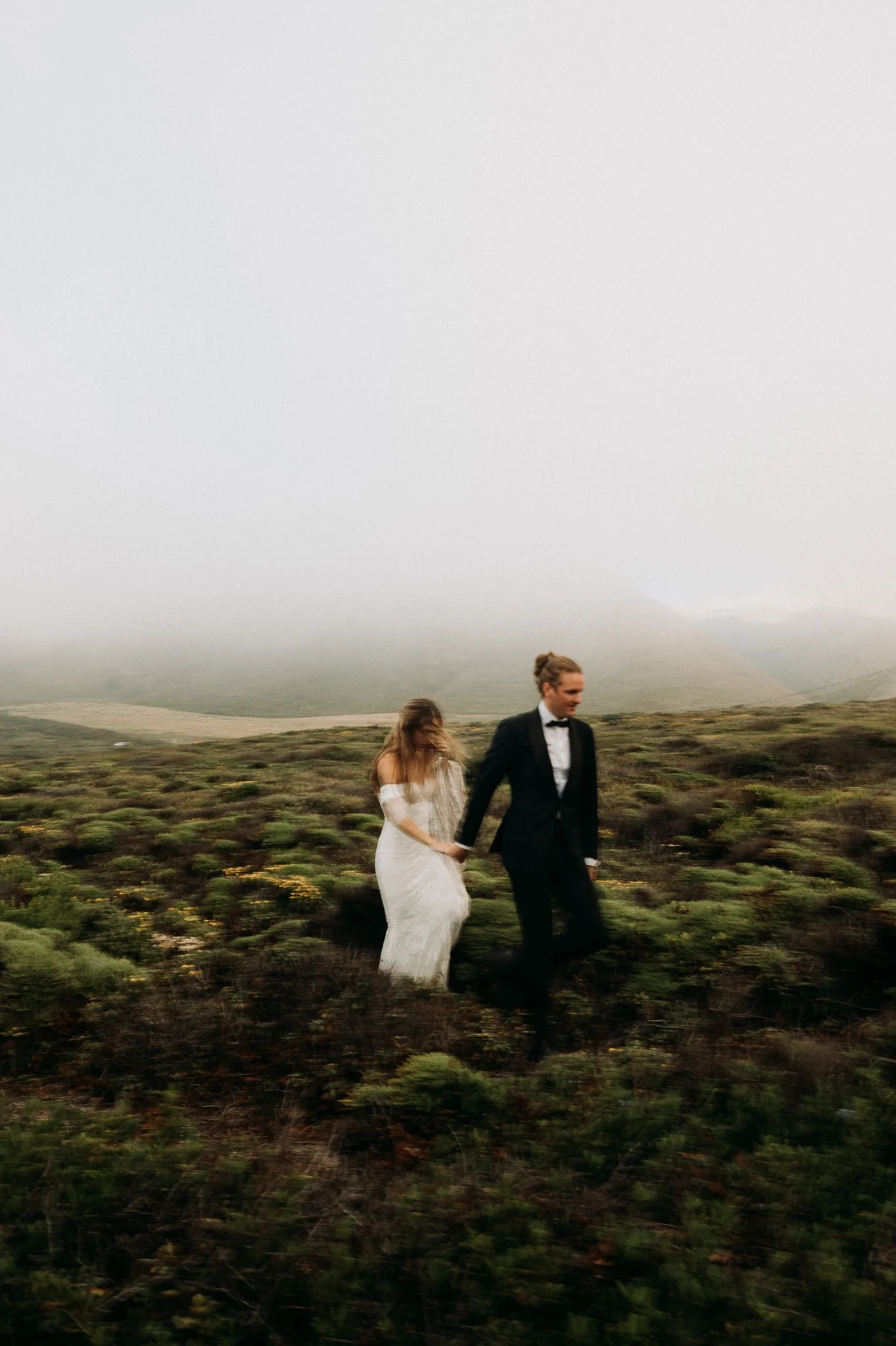A bride and groom walking hand in hand through a foggy, open landscape with green bushes and rolling hills.