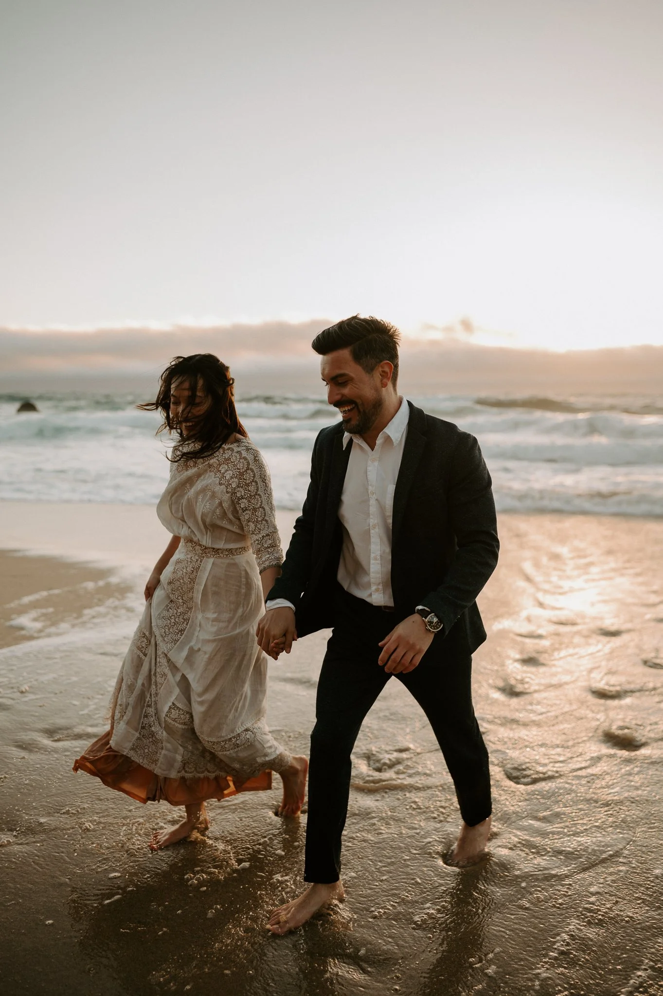 A couple walking barefoot in the ocean at sunset, holding hands and smiling.