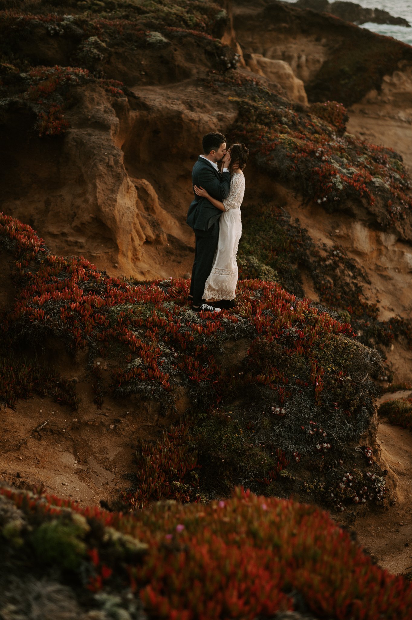A couple in wedding attire sharing a kiss on a cliffside surrounded by red and green desert plants with rocky cliffs and ocean in the background.