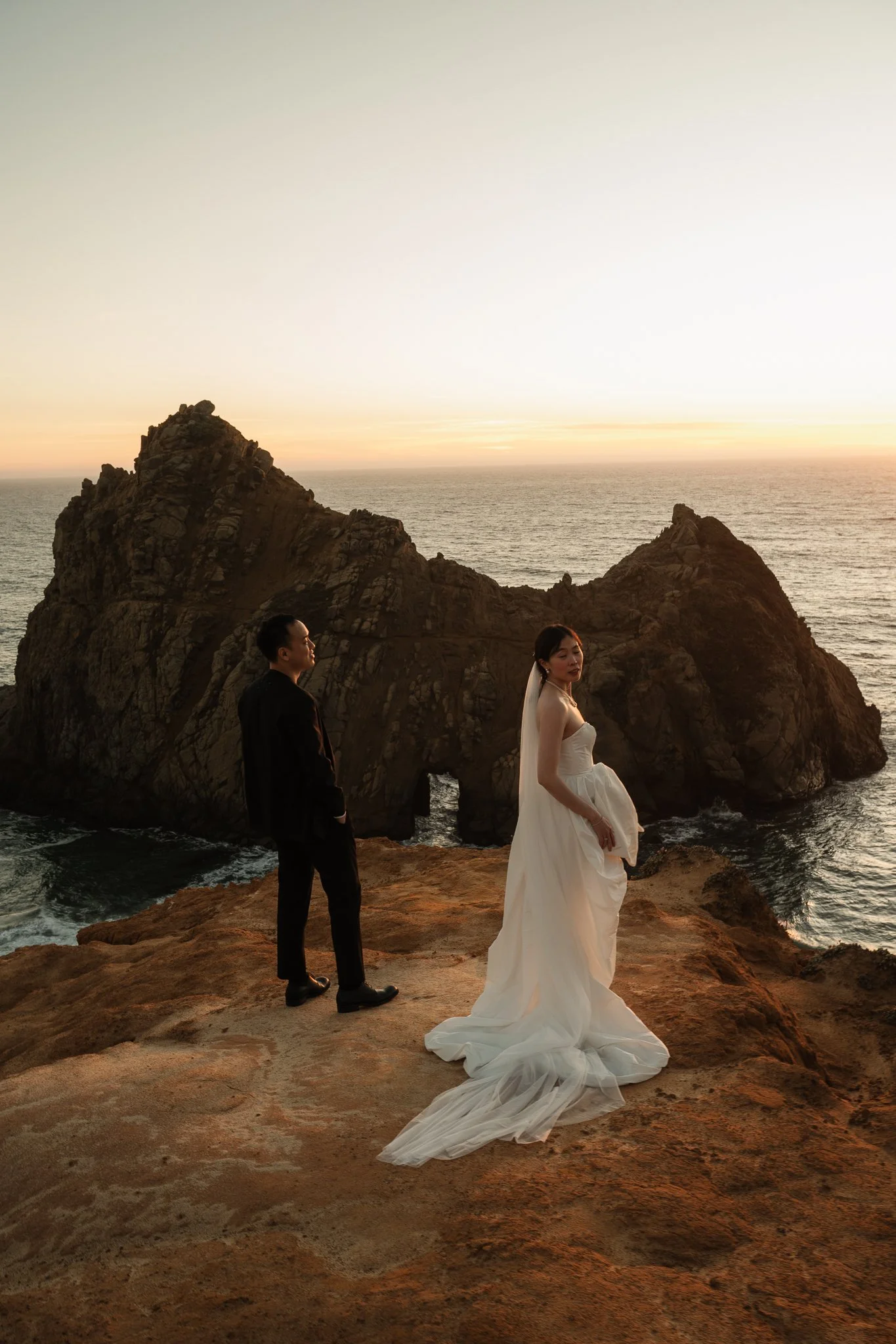 A bride and groom stand on a rocky cliffside overlooking the ocean at sunset, with a large rock formation in the background.