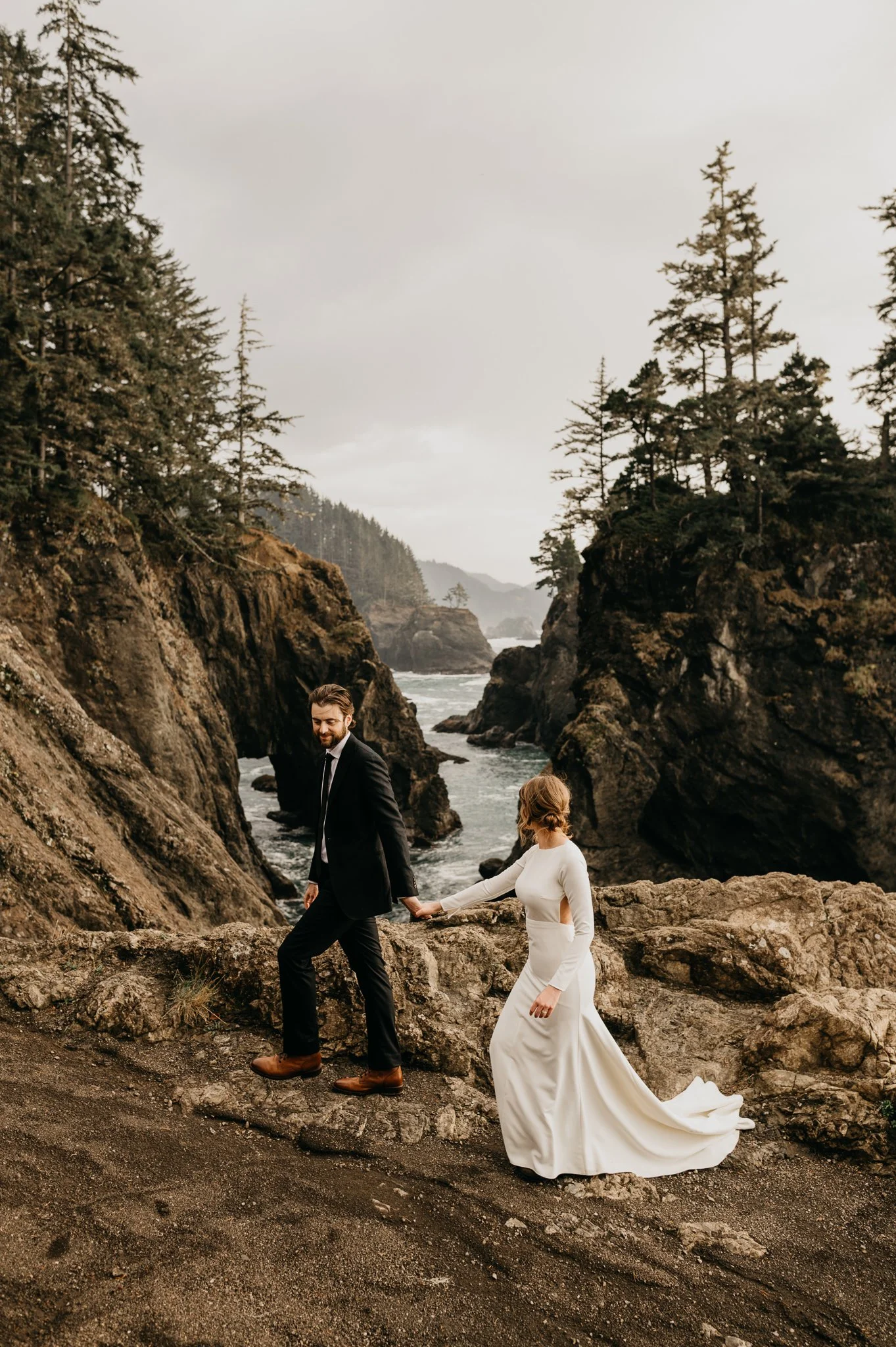 A bride and groom walking hand in hand on a rocky coastal trail with cliffs, trees, and the ocean in the background.