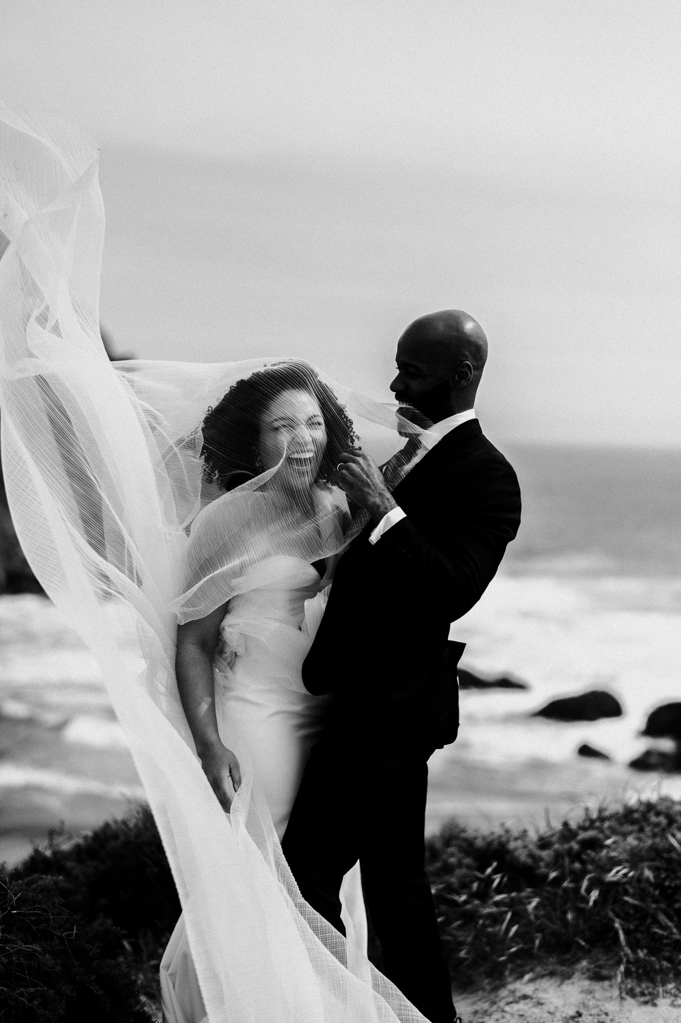 A couple in wedding attire sharing a joyful moment on a beach, with the bride laughing and the groom smiling, surrounded by a flowing veil.