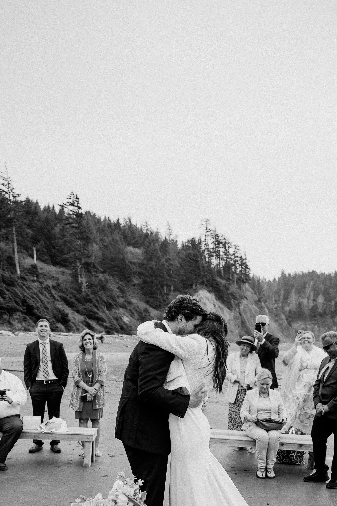A black and white photo of a wedding kiss on a beach, with guests smiling and taking pictures in the background, surrounded by trees and cliffs.