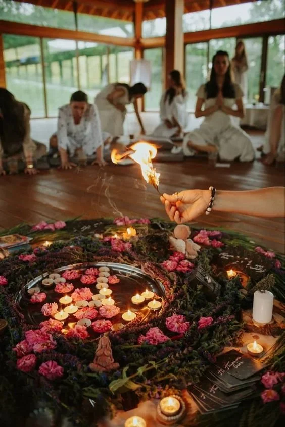 Indoor ritual circle with candles, flowers, incense, and people meditating in a wooden room.