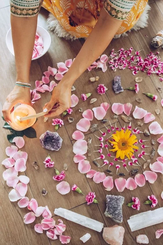 Flatlay of a spiritual altar with flowers, crystals, and a candle on a wooden floor. A person is holding a lit match near the candle.
