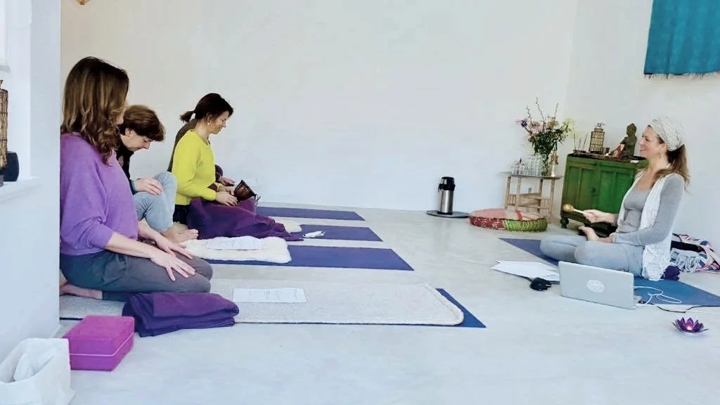 A yoga class with a group of individuals seated on mats in a meditation pose, facing an instructor. The room has minimalist decor with soft lighting, flowers, and a small cabinet in the background. A laptop and a few personal items are placed on the floor in front of the instructor.