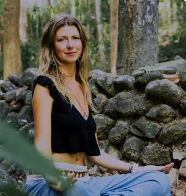 Person sitting cross-legged outdoors near stone wall and trees, wearing black top and blue pants. Forest setting with greenery in background.