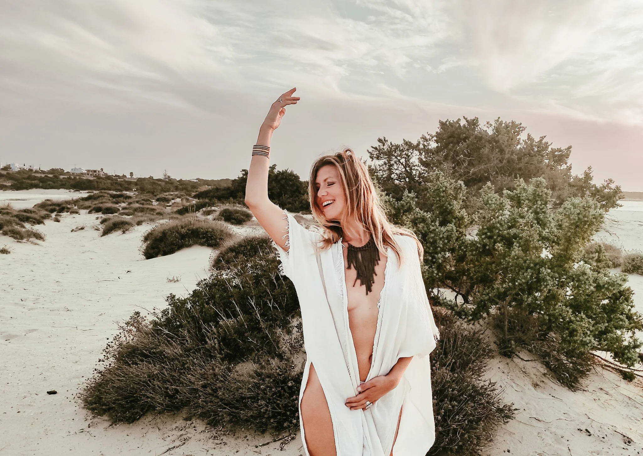 Woman in white robe posing on a sandy beach with bushes, wearing jewelry, and smiling.