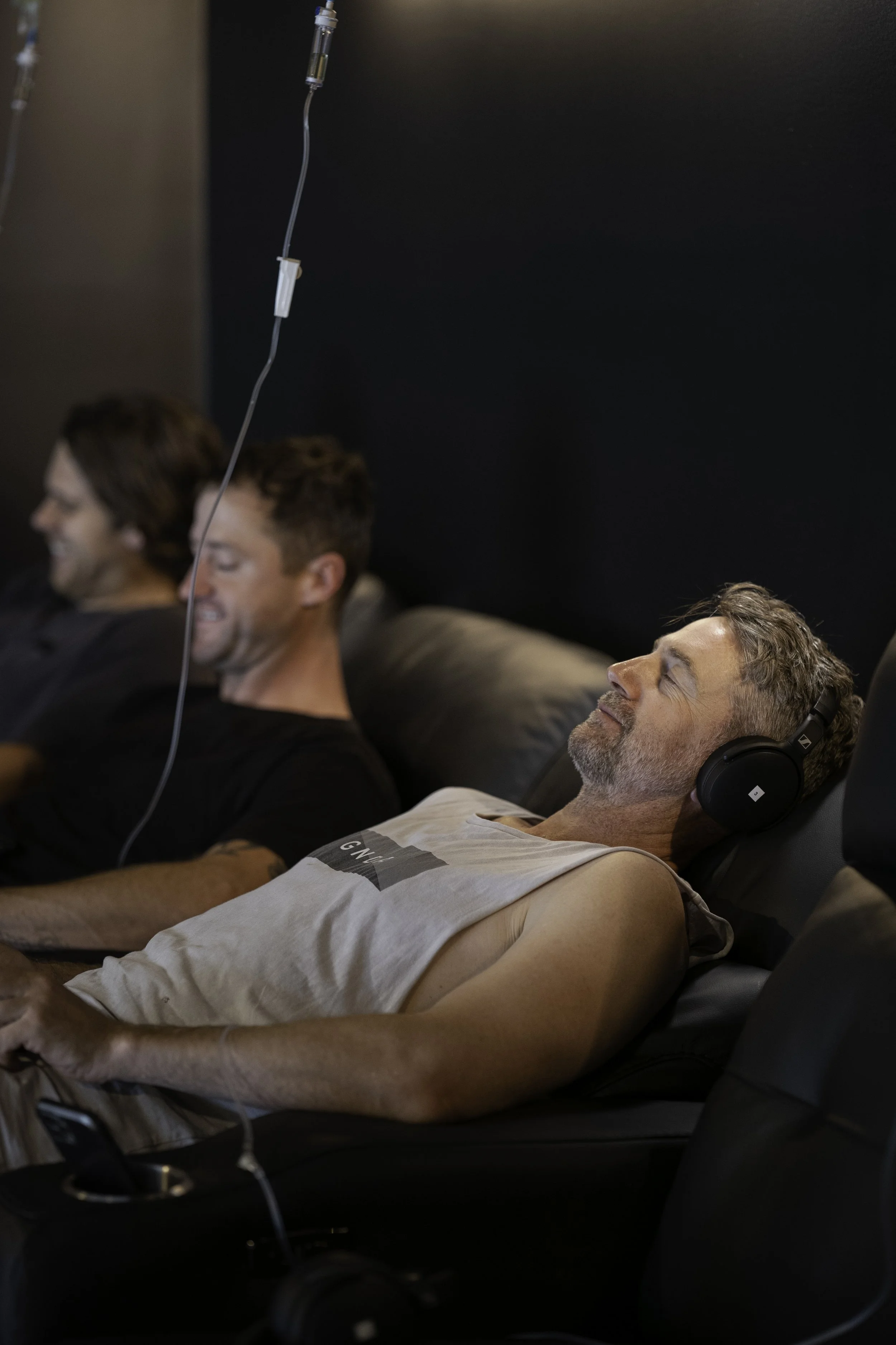 Three men are sitting on couches, smiling and relaxing. The man in the foreground is lying back with eyes closed, wearing headphones, a tank top, and has an IV inserted into his arm.