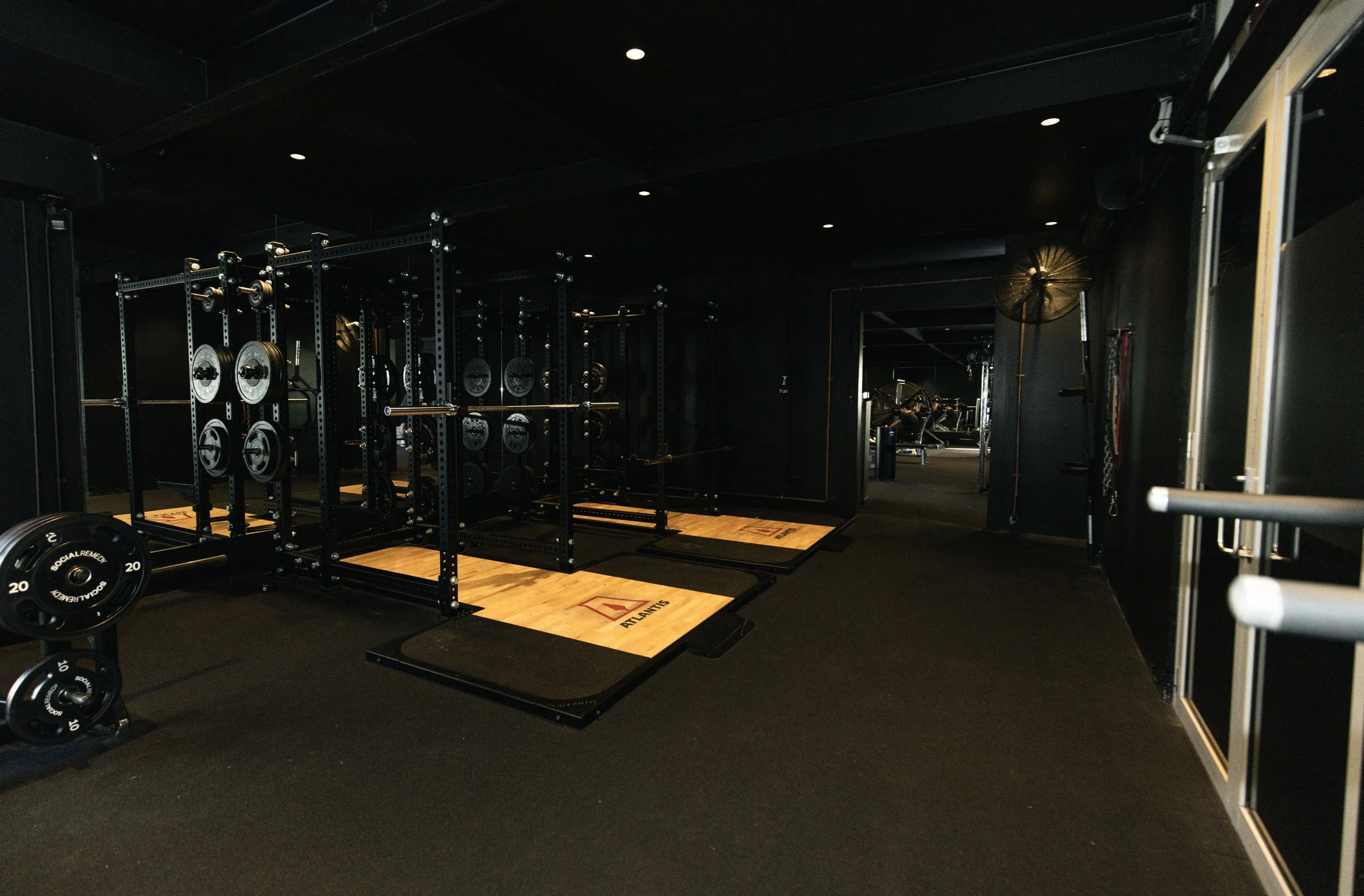 A black gym with strength training equipment, including Atlantis squat racks with weights and barbell plates, wooden platforms for lifting, and a mirror on the right wall. The room has a black ceiling, dark walls, and overhead lighting.