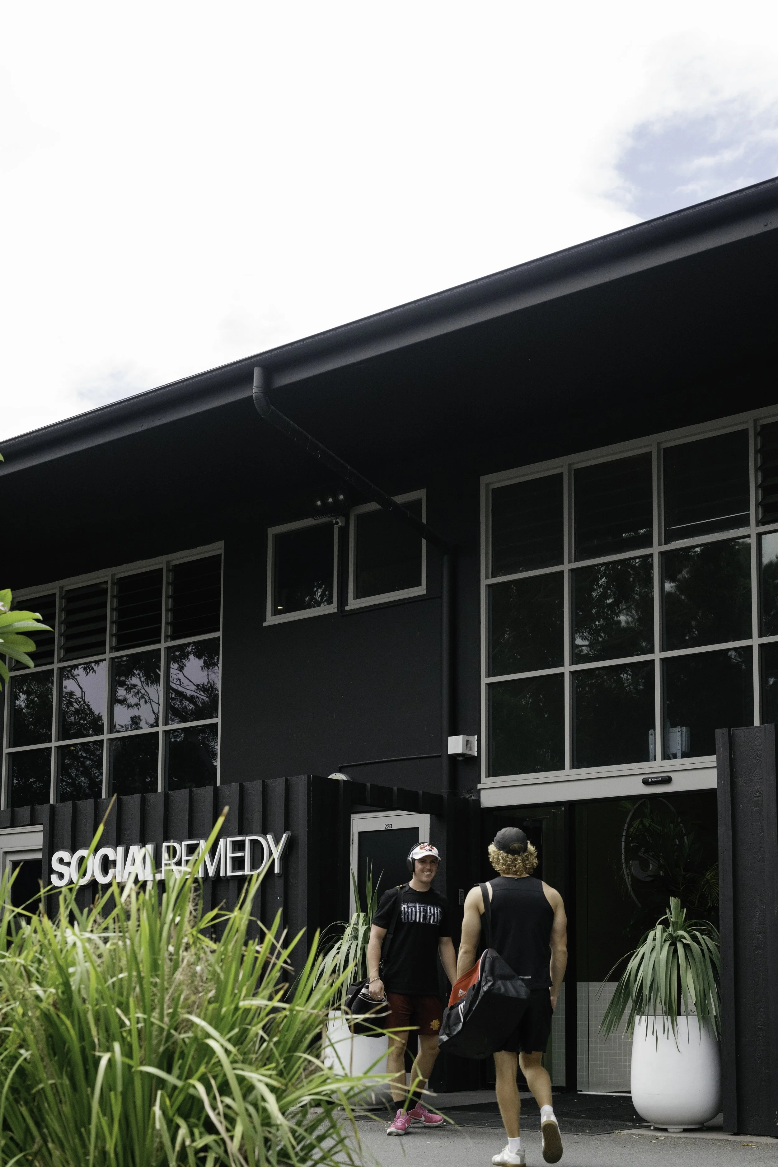 Two men standing and talking outside a modern building with large glass windows and dark exterior, with a sign that reads 'Social Remedy'. One  is smiling, wearing a black T-shirt and maroon shorts, and the other has curly hair, wearing black shorts and a black tank top, carrying a backpack. There are large potted plants and greenery in the foreground.