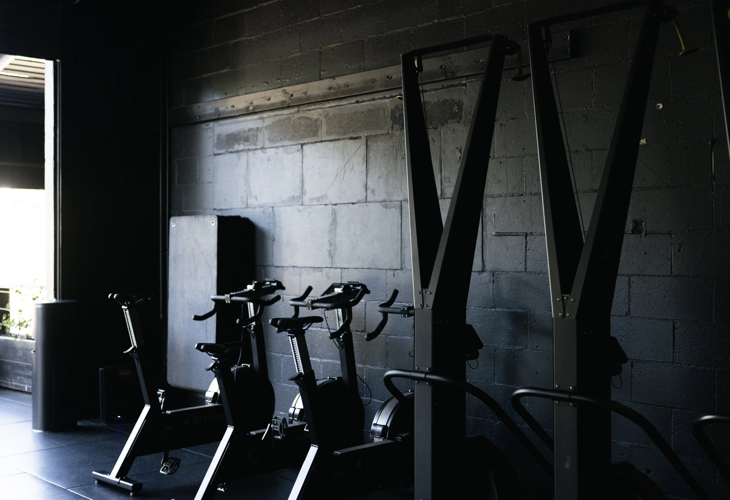 An indoor gym area with stationary bikes and weightlifting equipment against a dark brick wall, lit by natural light from an open door.