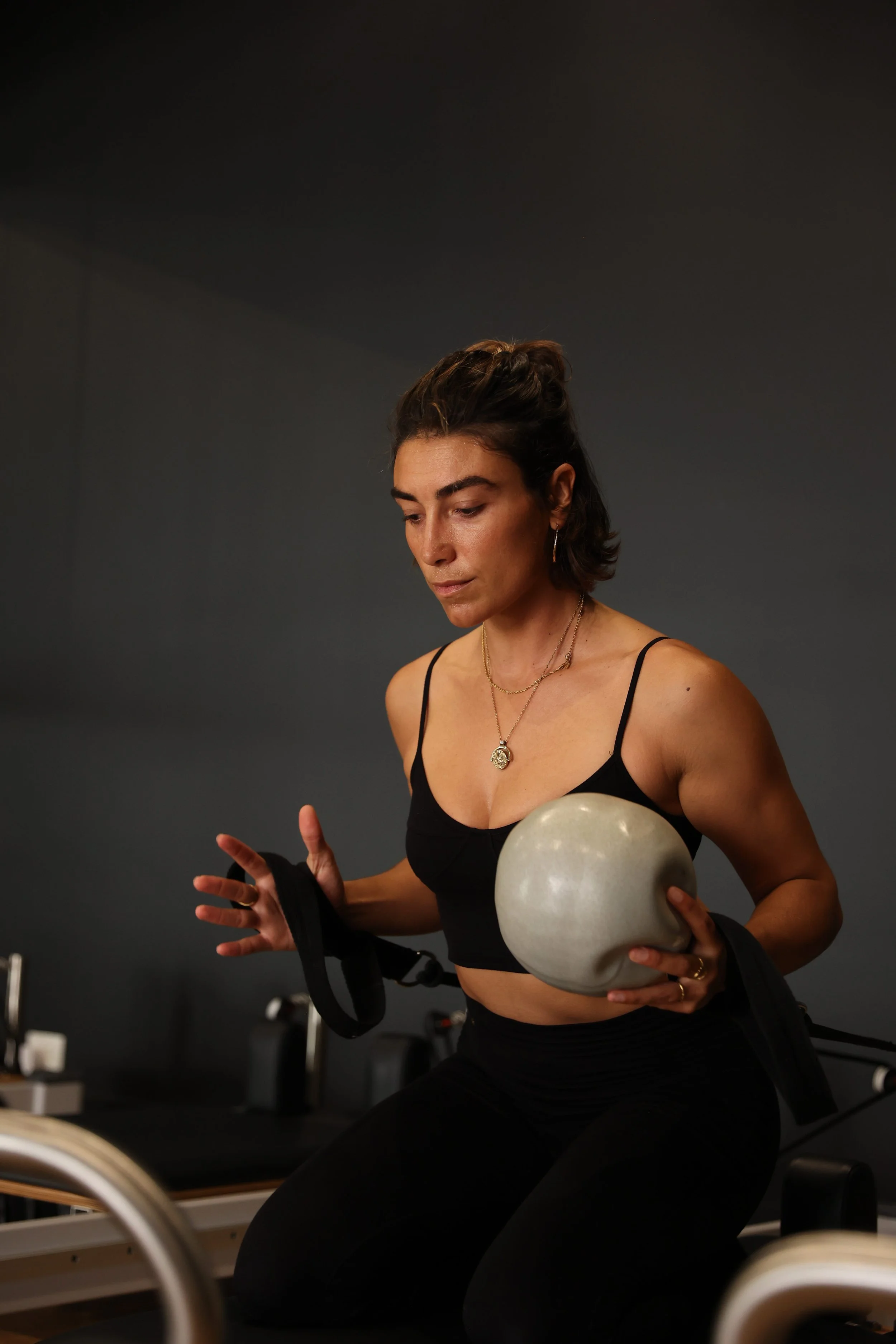 A woman in athletic attire holding a light-colored ball on a reformer machine, with reformers in the background.