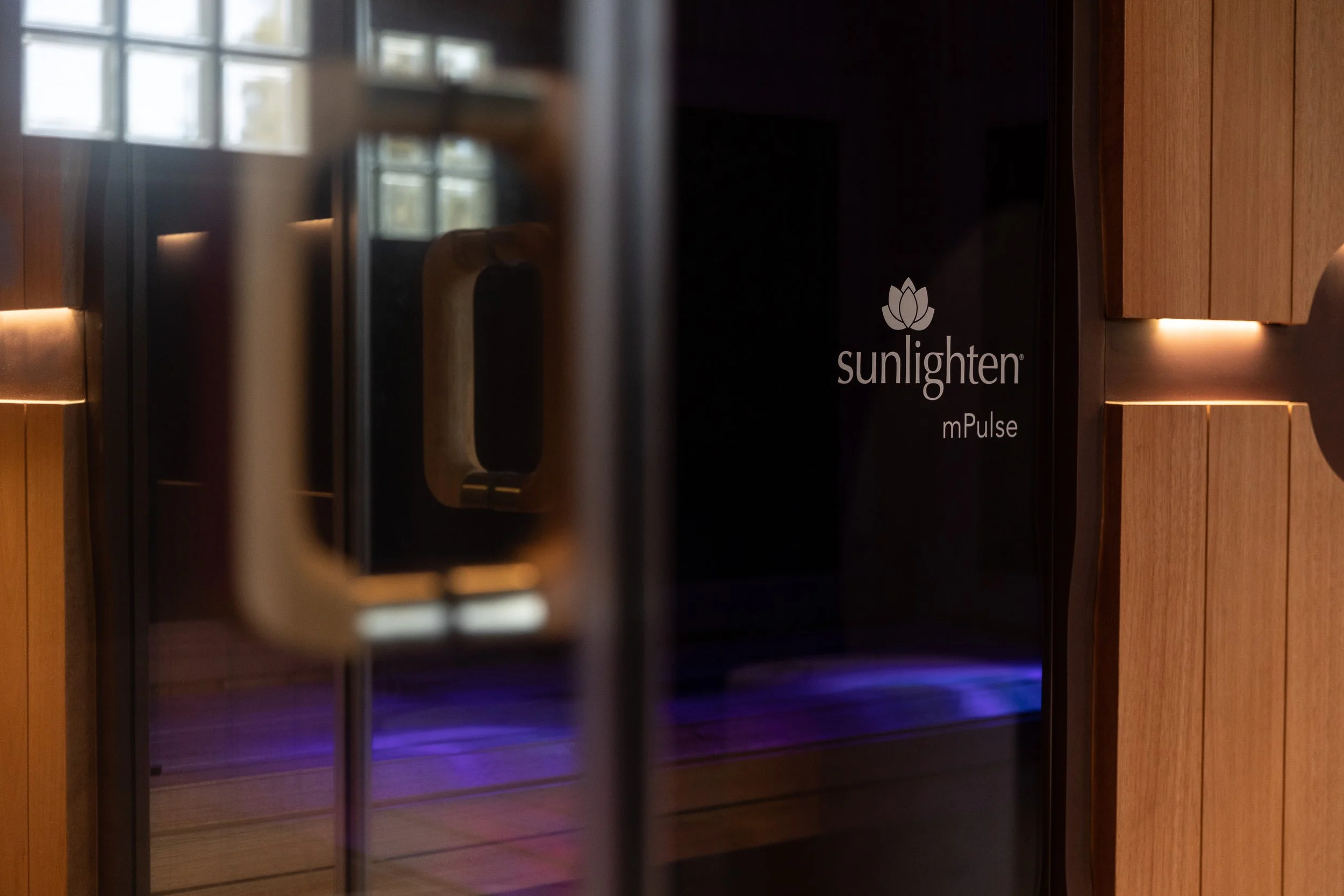 A black Sunlighten mPulse sauna with wooden accents and a door handle, reflecting a window and LED lighting inside.