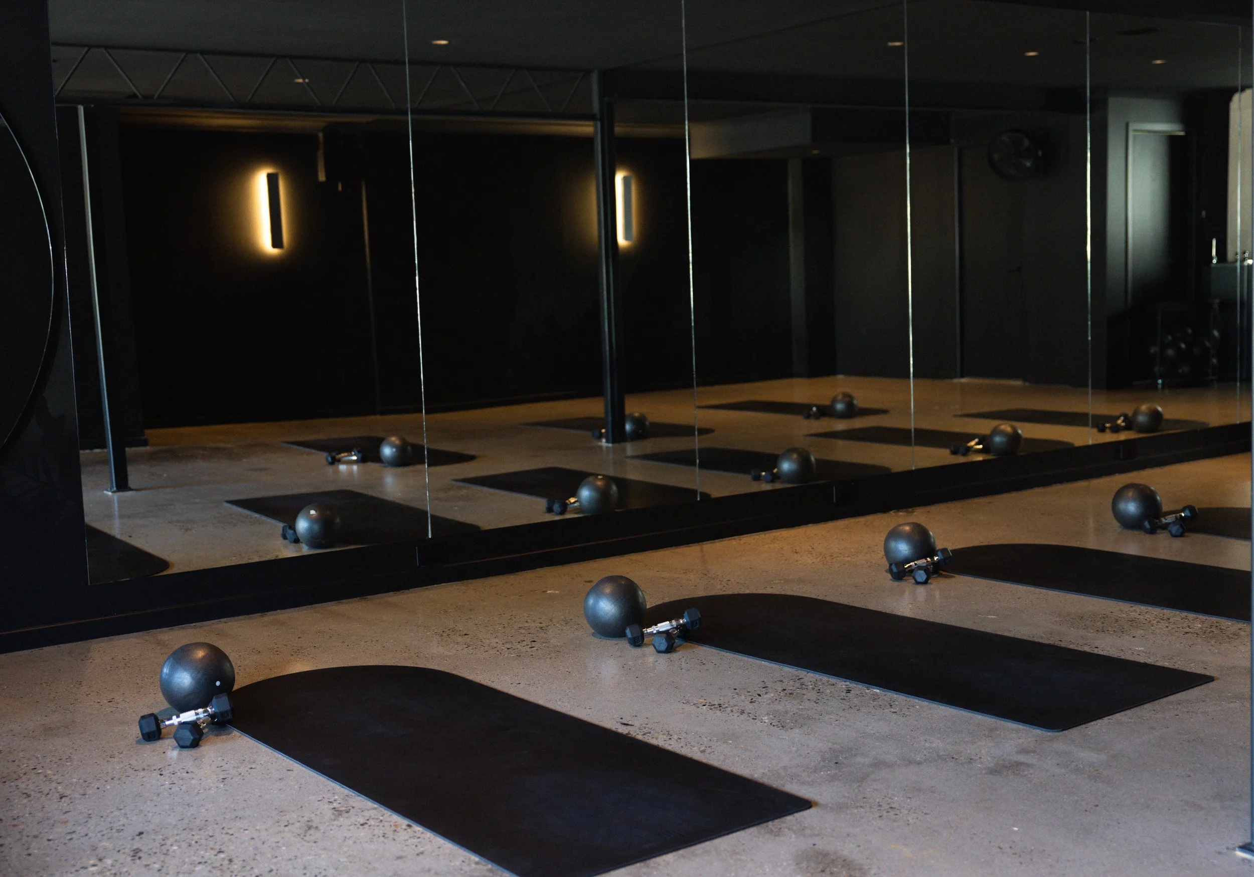 Fitness studio with black mats and exercise balls arranged in front of mirrors, dim lighting, and a few wall lights in the background.