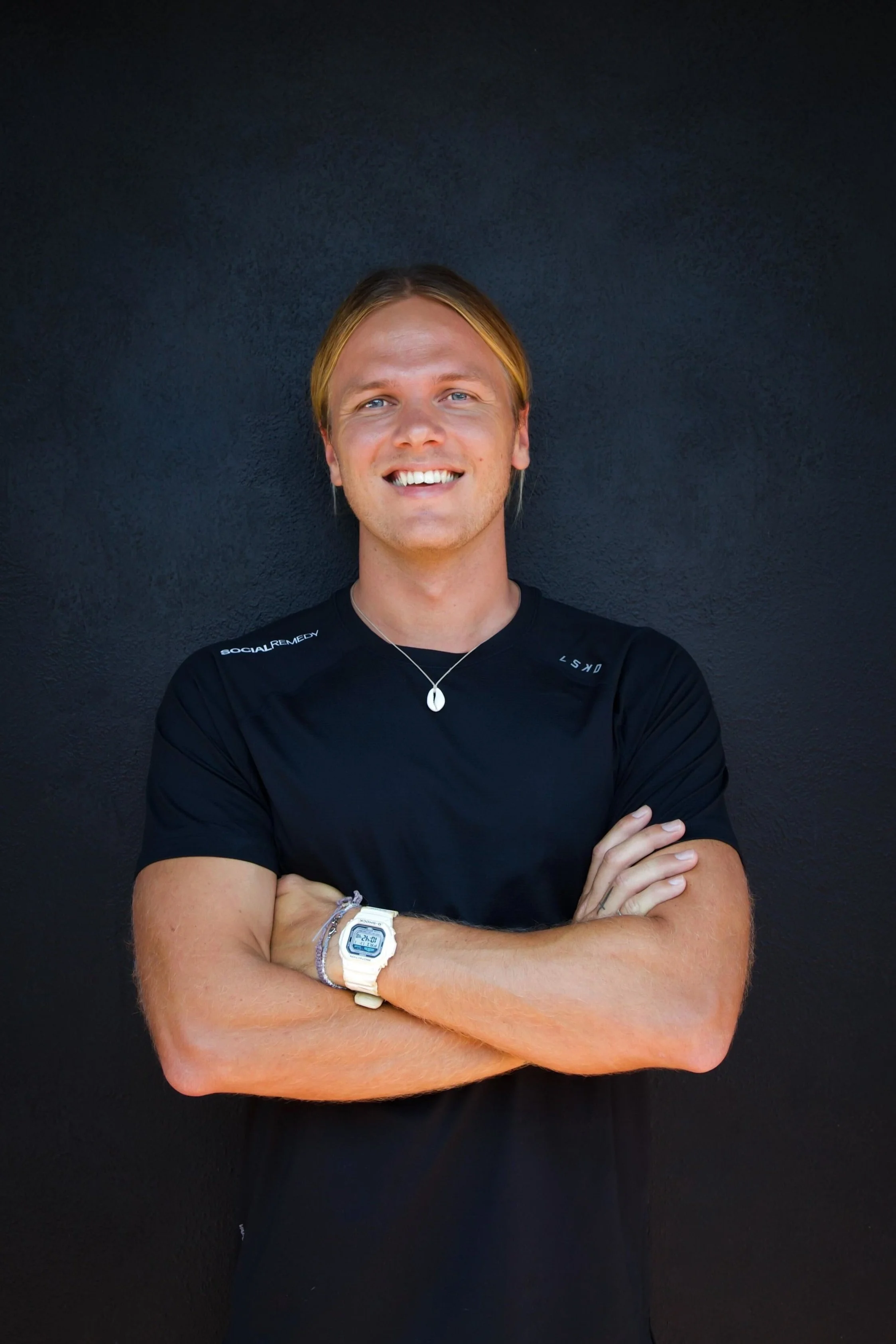 A young man with long blonde hair smiling against a black background, wearing a black athletic t-shirt, a white digital watch, a silver necklace, and a bracelet.