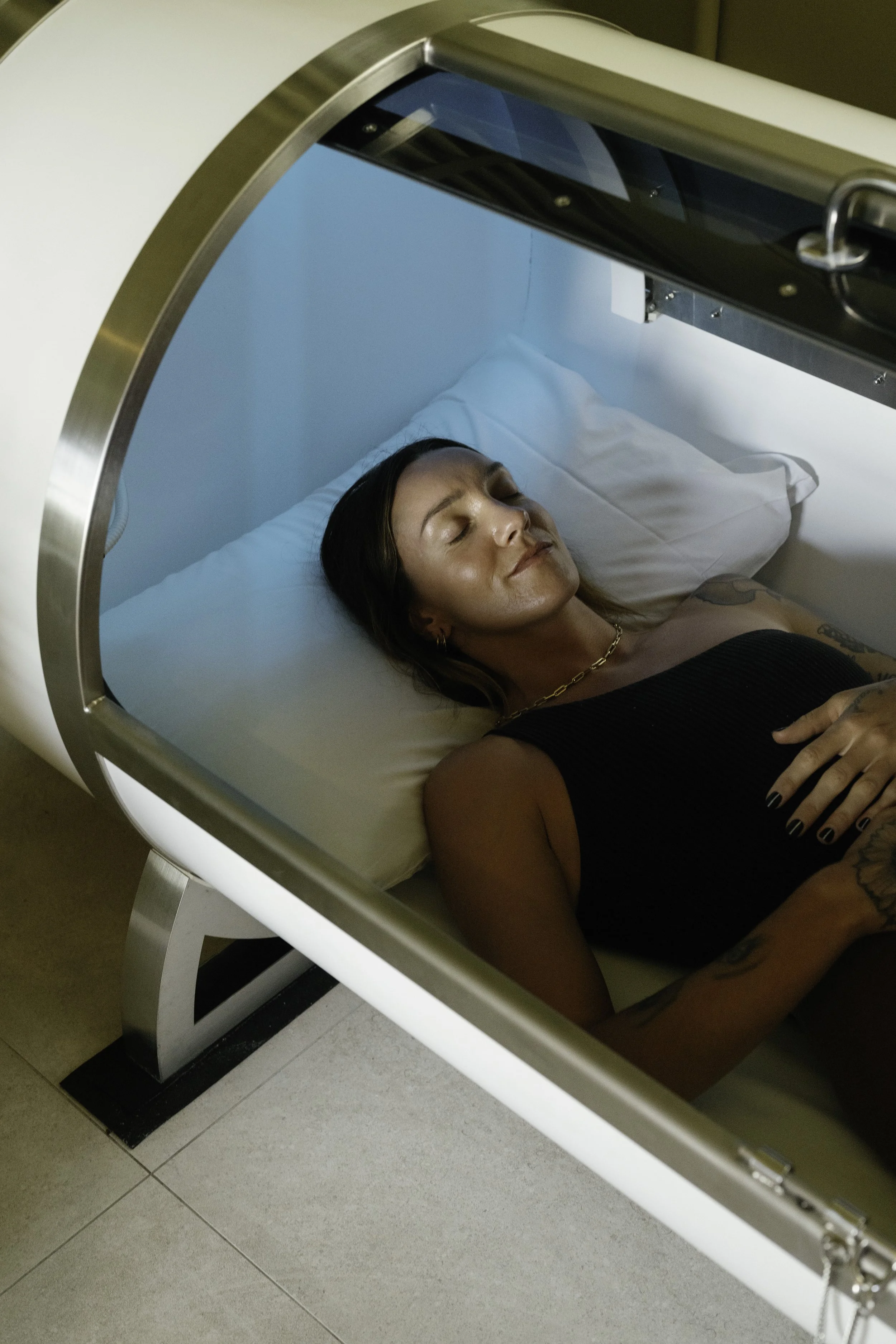 A woman with dark hair and tattoos lying inside a Hyperbaric Oxygen Therapy chamber, appearing to be in a relaxed or sleeping state.