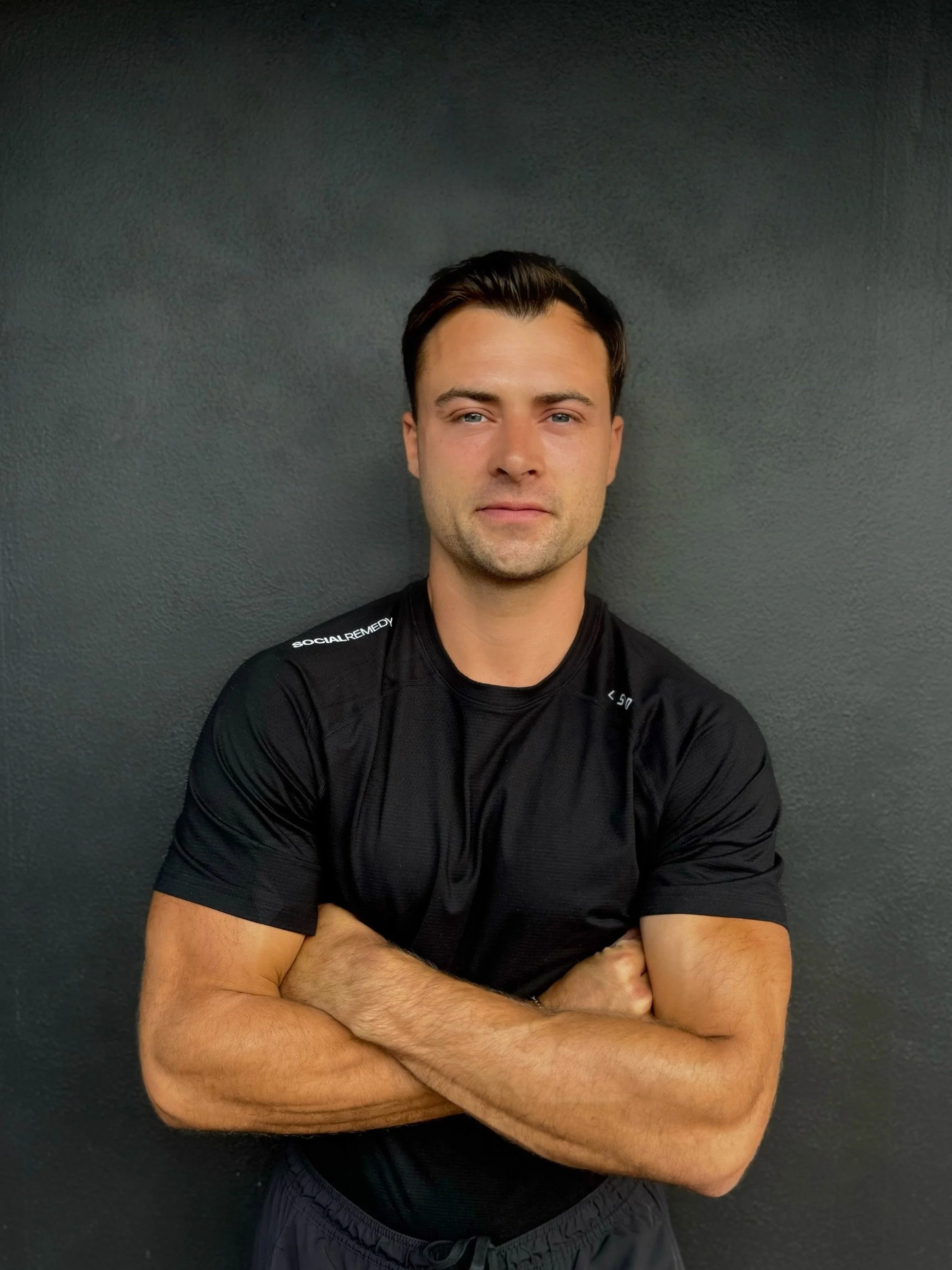 A young man with dark hair and fair skin, wearing a black athletic shirt with white text on the shoulder, crossing his arms and standing against a dark gray wall.
