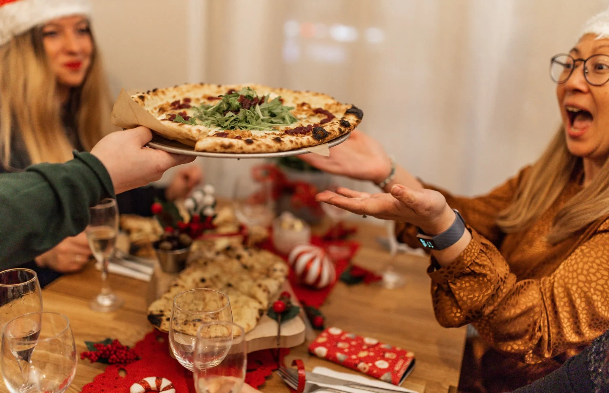 Person passing a pizza with greens to another person at a Christmas celebration table.