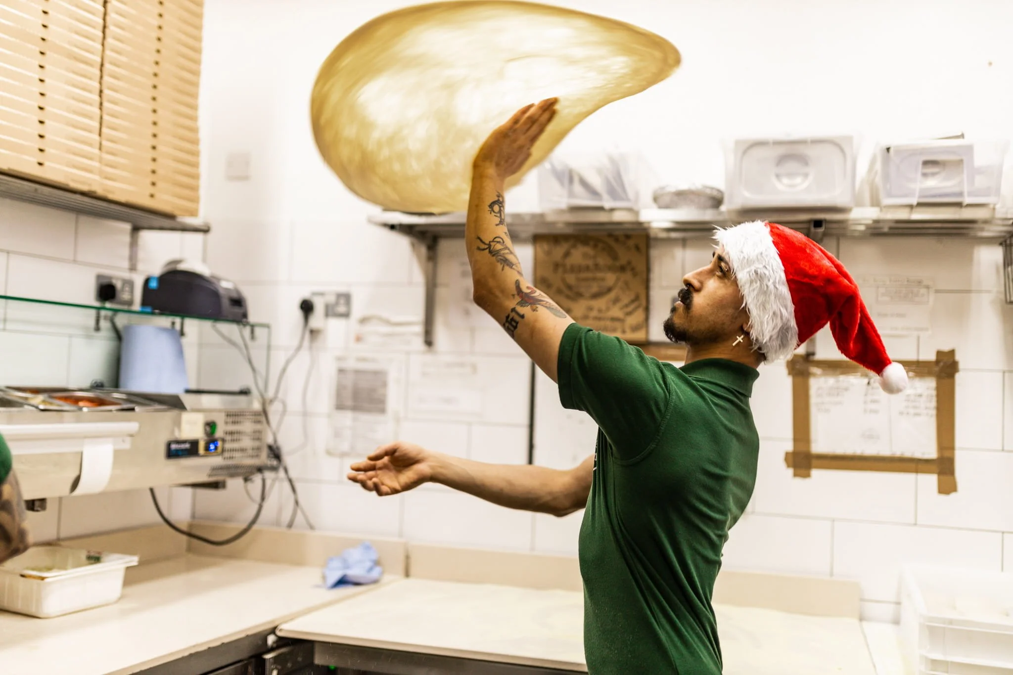 A man wearing a Santa hat raises a pizza dough in a kitchen, preparing for baking.