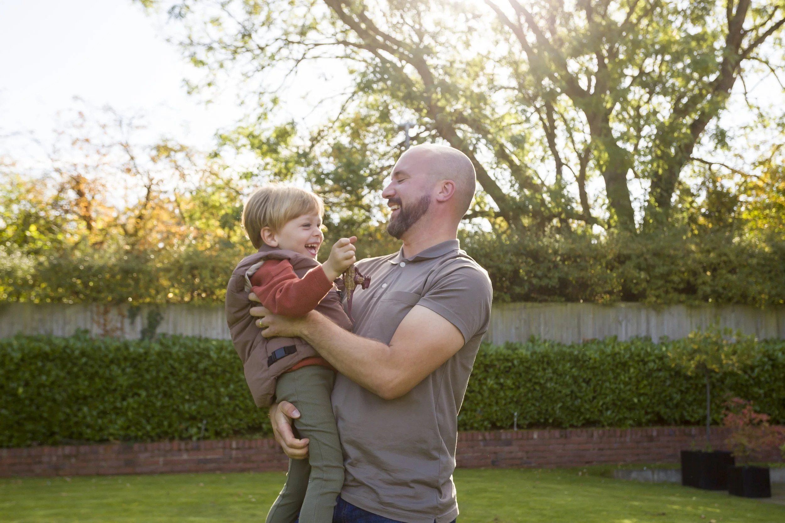 A man holding a young boy outdoors in front of a wooden fence and green bushes, with sunlight shining through trees in the background.