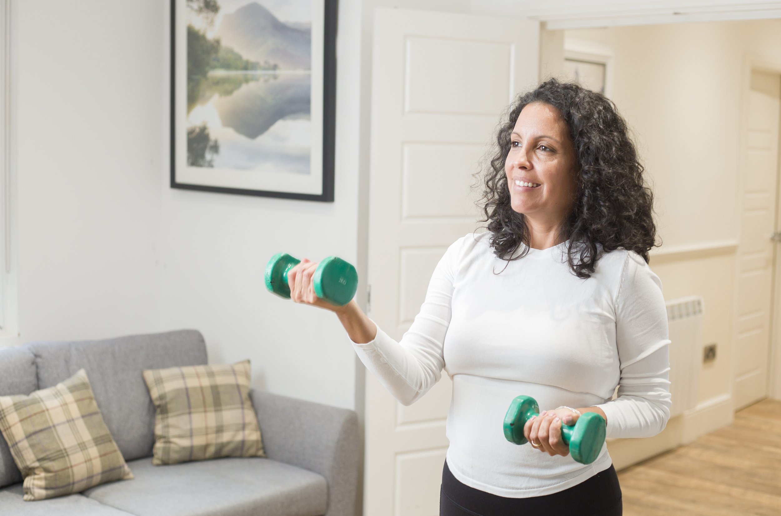 A woman with curly dark hair wearing a white long-sleeve shirt and black pants, exercising with green dumbbells in a living room decorated with a gray sofa, plaid pillows, and a painting of a mountain and lake scene on the wall.