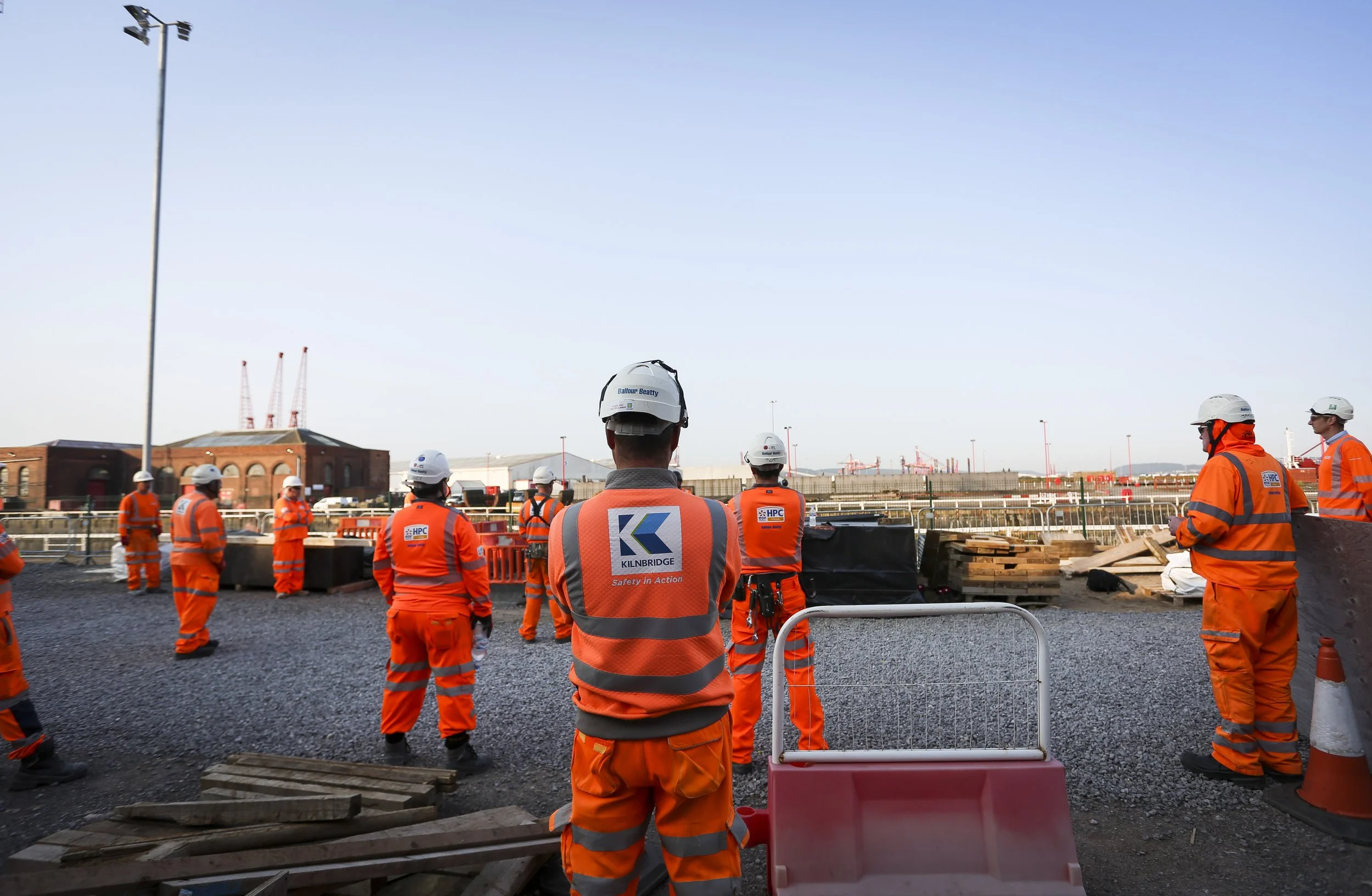 Construction workers wearing orange high-visibility clothing and safety helmets gathered on a construction site, with industrial buildings, cranes, and a clear sky in the background.