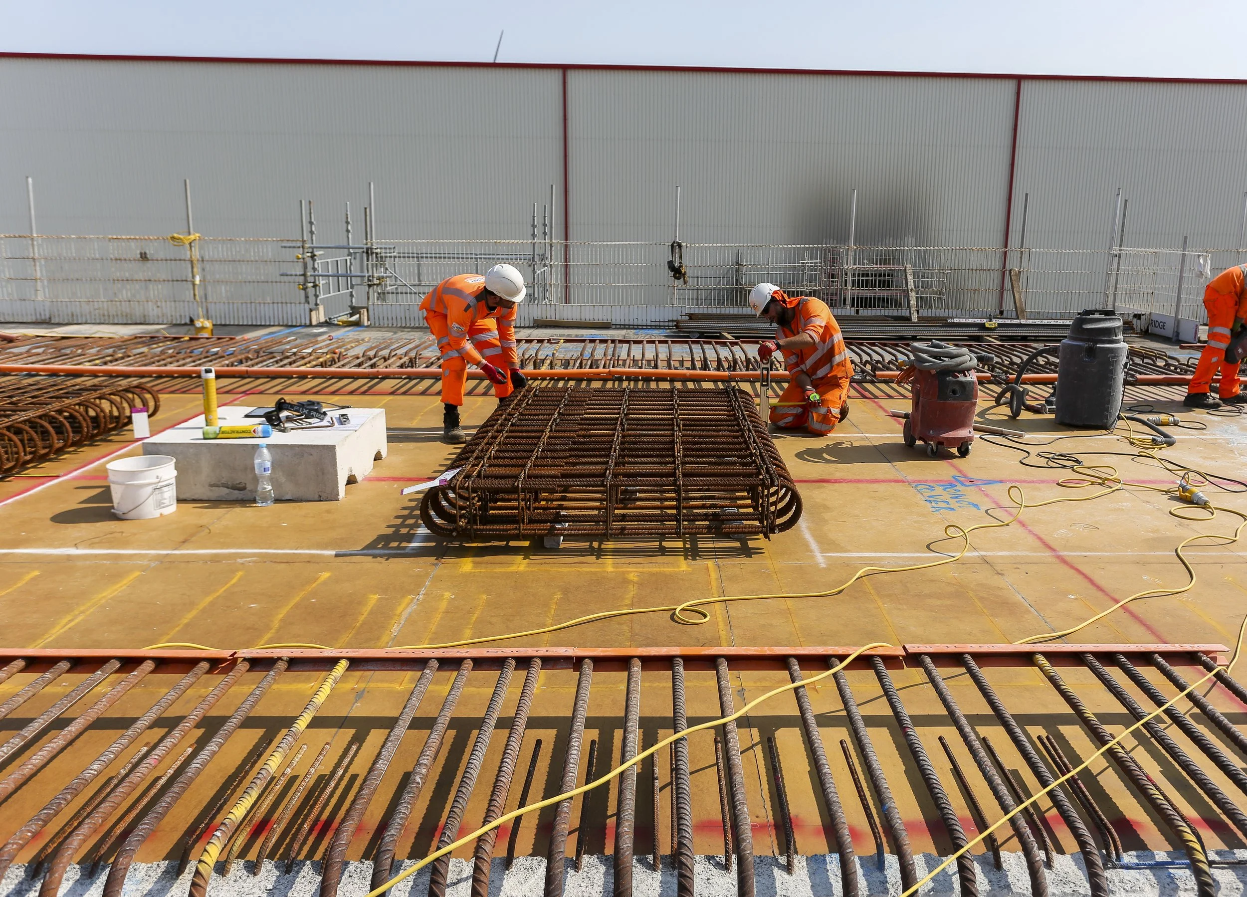 Construction workers wearing safety helmets and orange safety vests working on rebar reinforcement for a building. They are on a partially built structure with tools, equipment, and wires around.