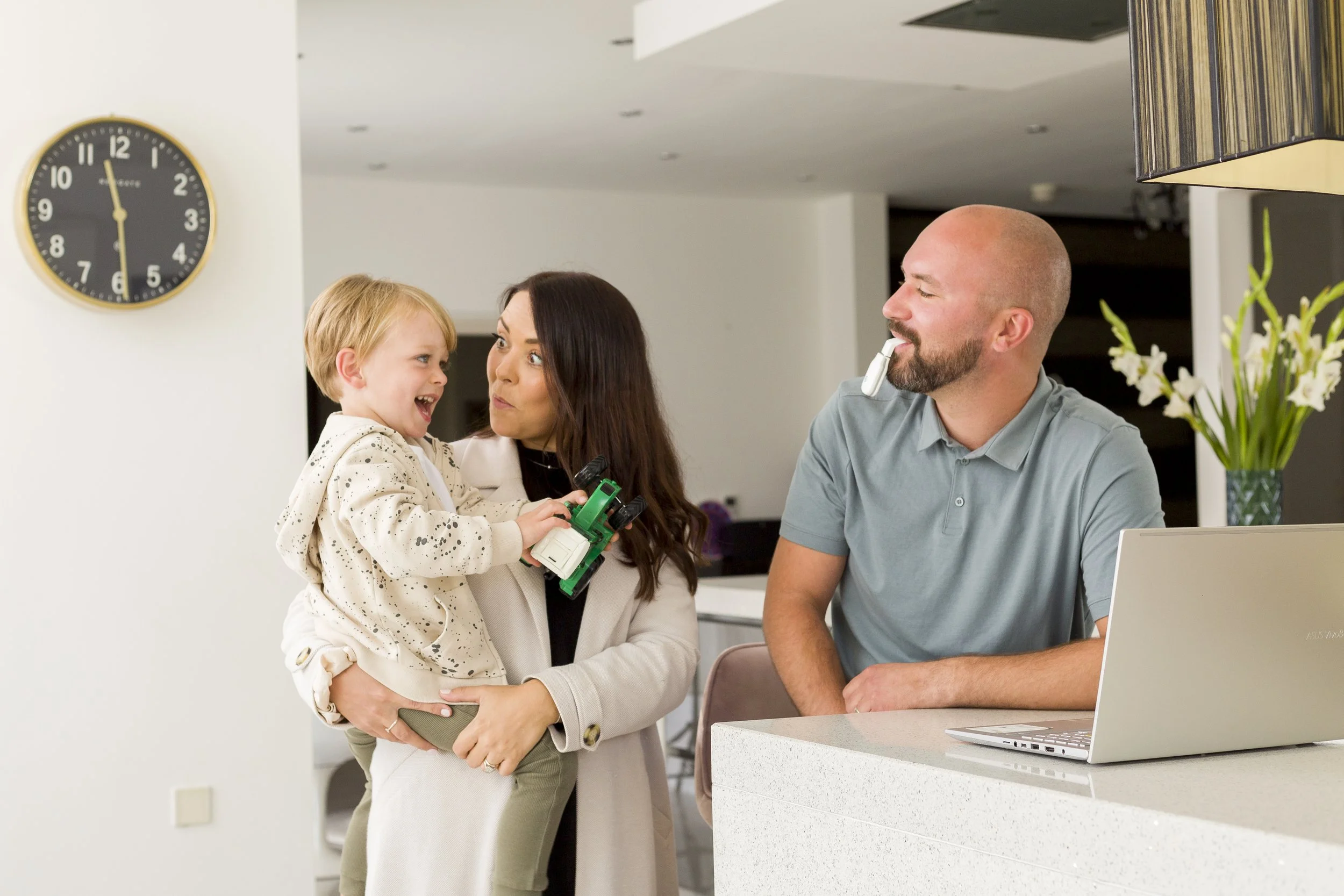 A woman holding a young boy while talking to a man at a reception desk in a modern building. The woman and boy are smiling; the boy is holding a toy car. The man has a clapperboard in his mouth and is dressed casually. A large wall clock shows approx