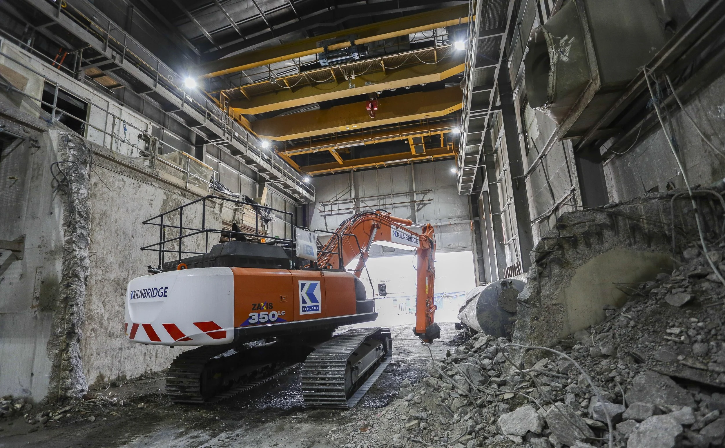Indoor construction site with an orange excavator moving debris, concrete walls, and running tracks on the machine, with construction equipment and scaffolding overhead.