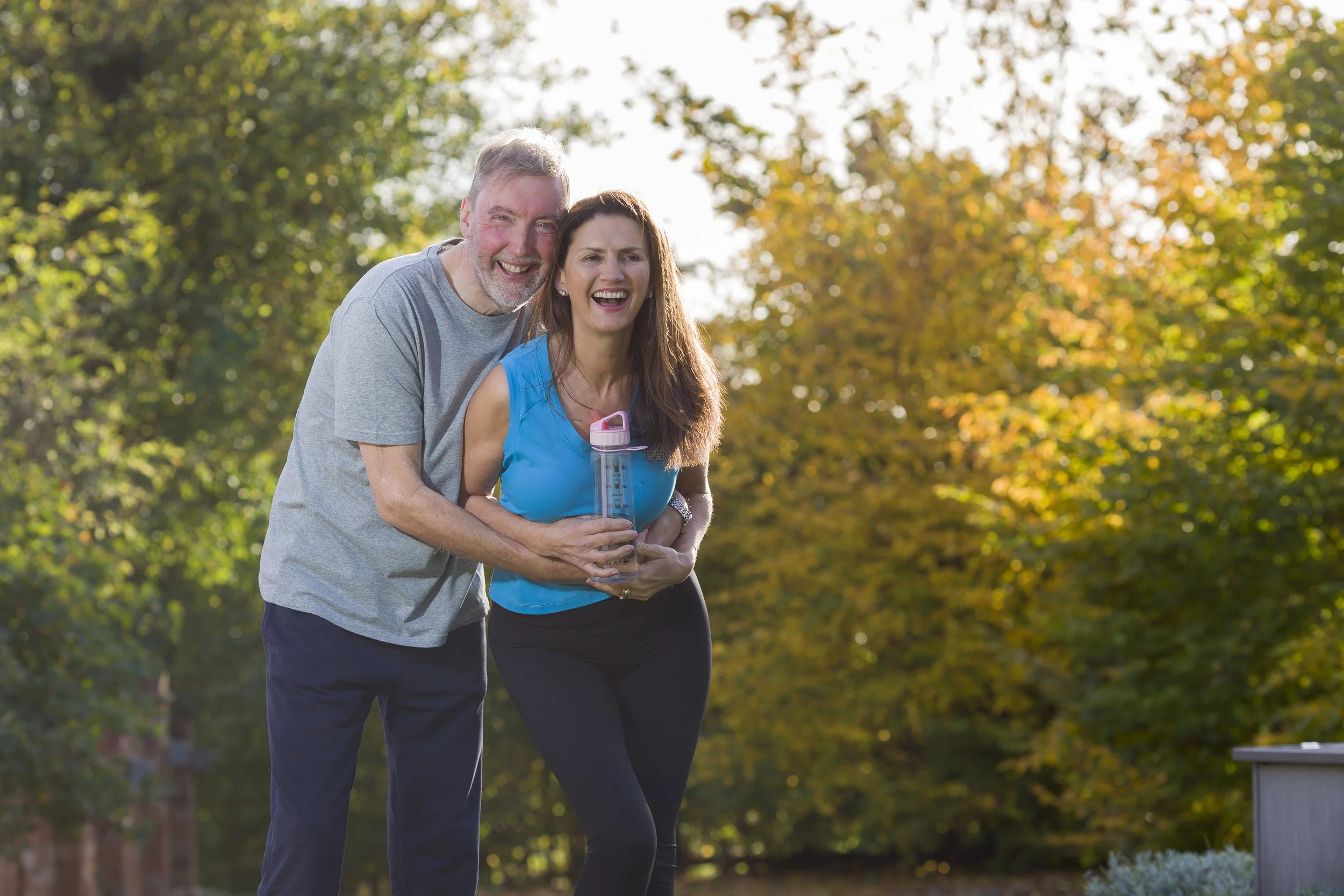 A happy couple with gray hair jogging outdoors in a park during fall, smiling and holding hands, with trees with yellow and green leaves in the background.