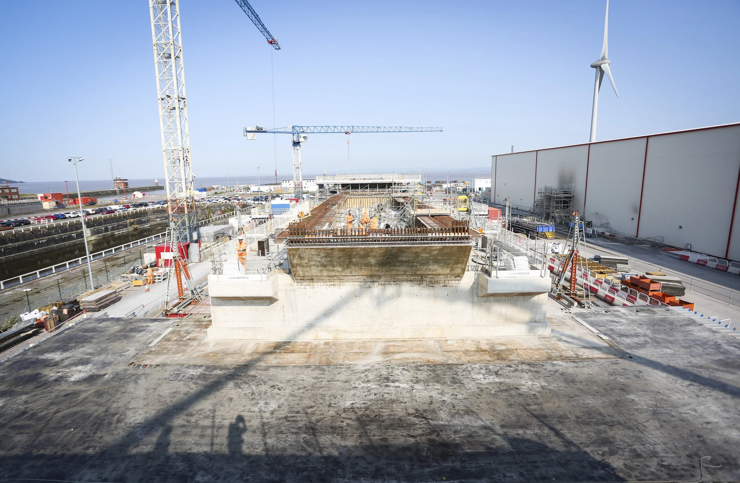Construction site with cranes, workers, and a partially built structure near a body of water, with a wind turbine in the background.