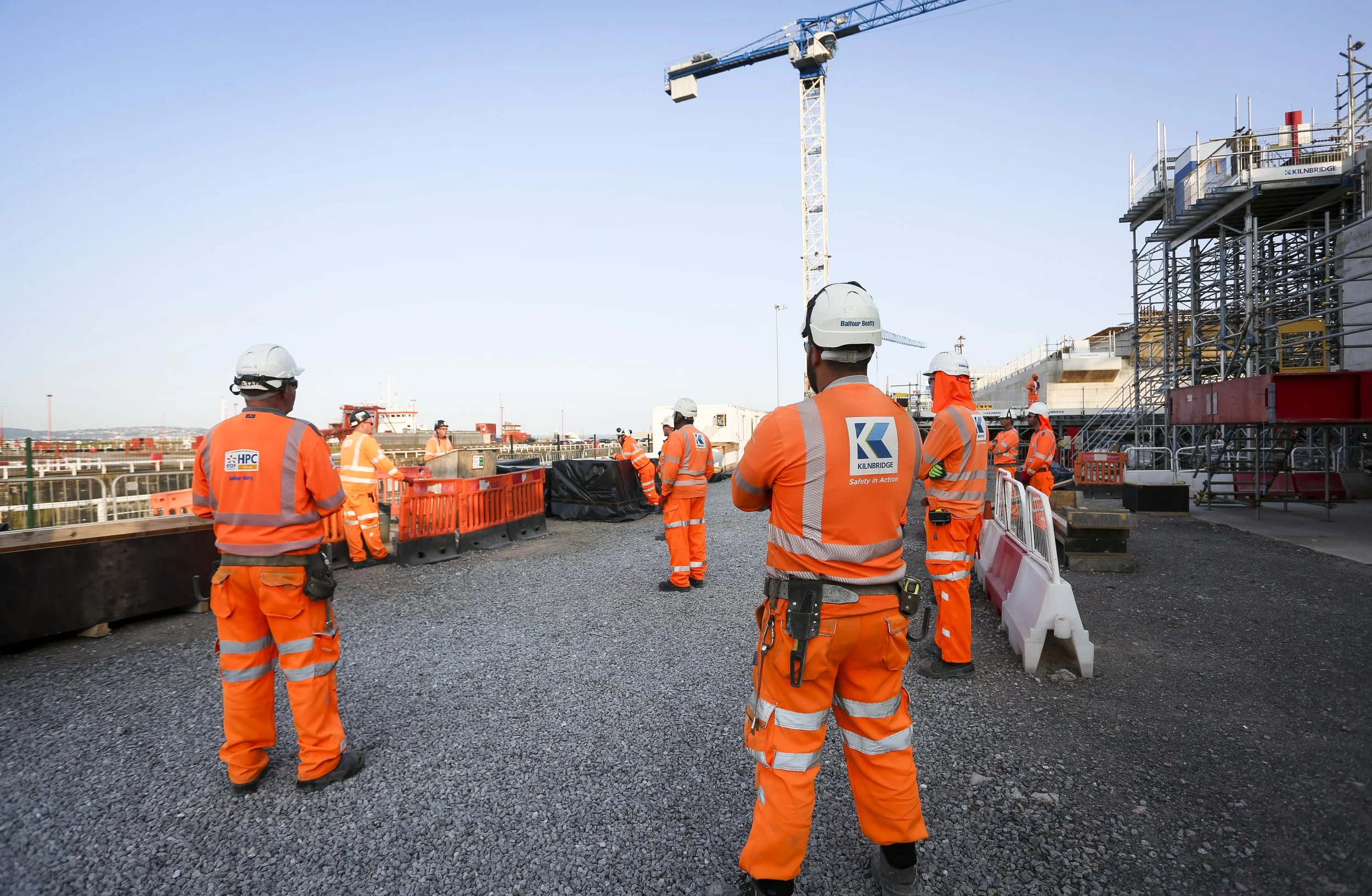 Construction workers in orange safety gear, including helmets and reflective vests, standing on a gravel surface at a construction site with scaffolding, barriers, and a crane in the background.