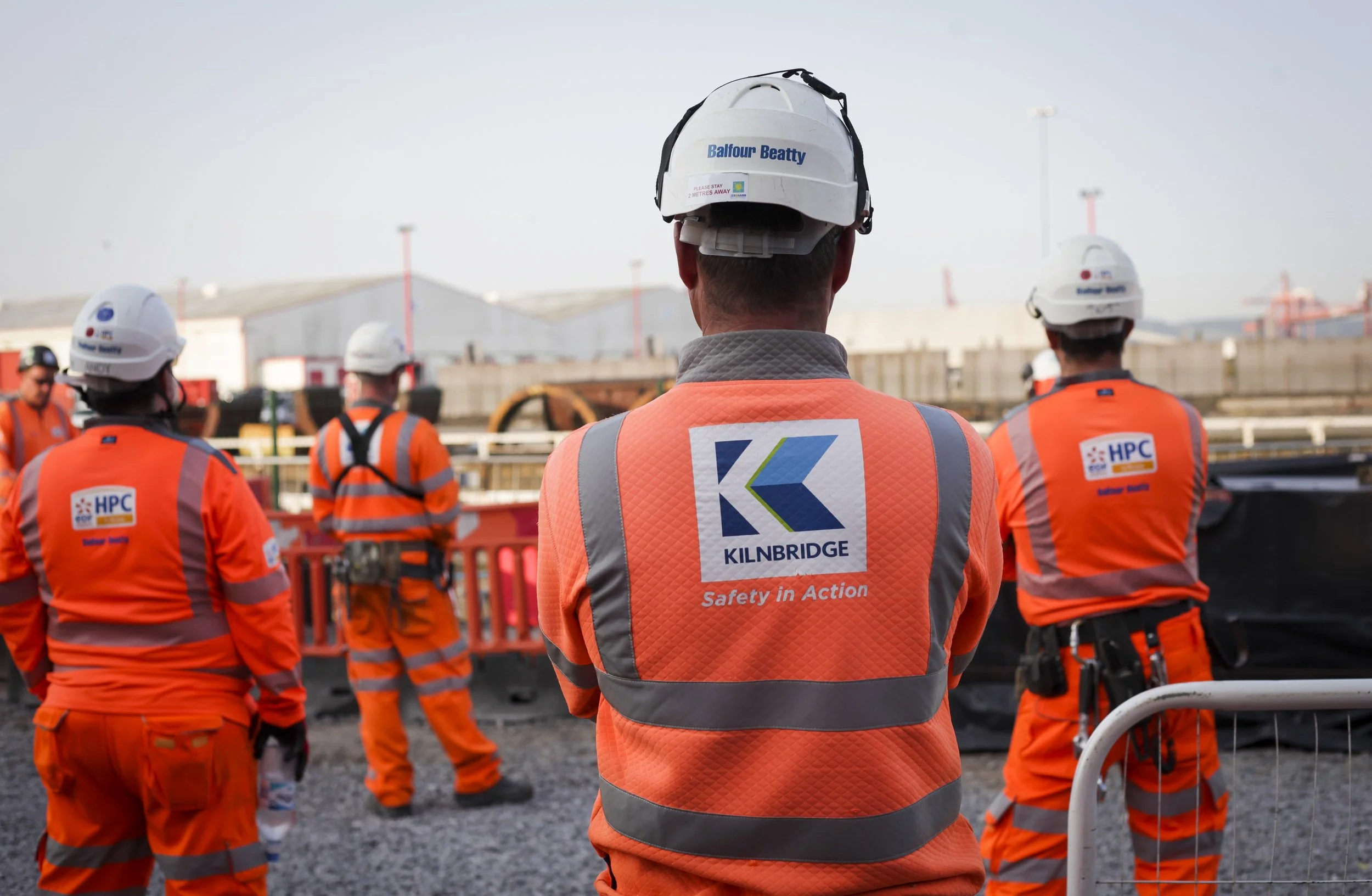 Construction workers in orange safety vests and white helmets standing at a construction site, with industrial buildings in the background.