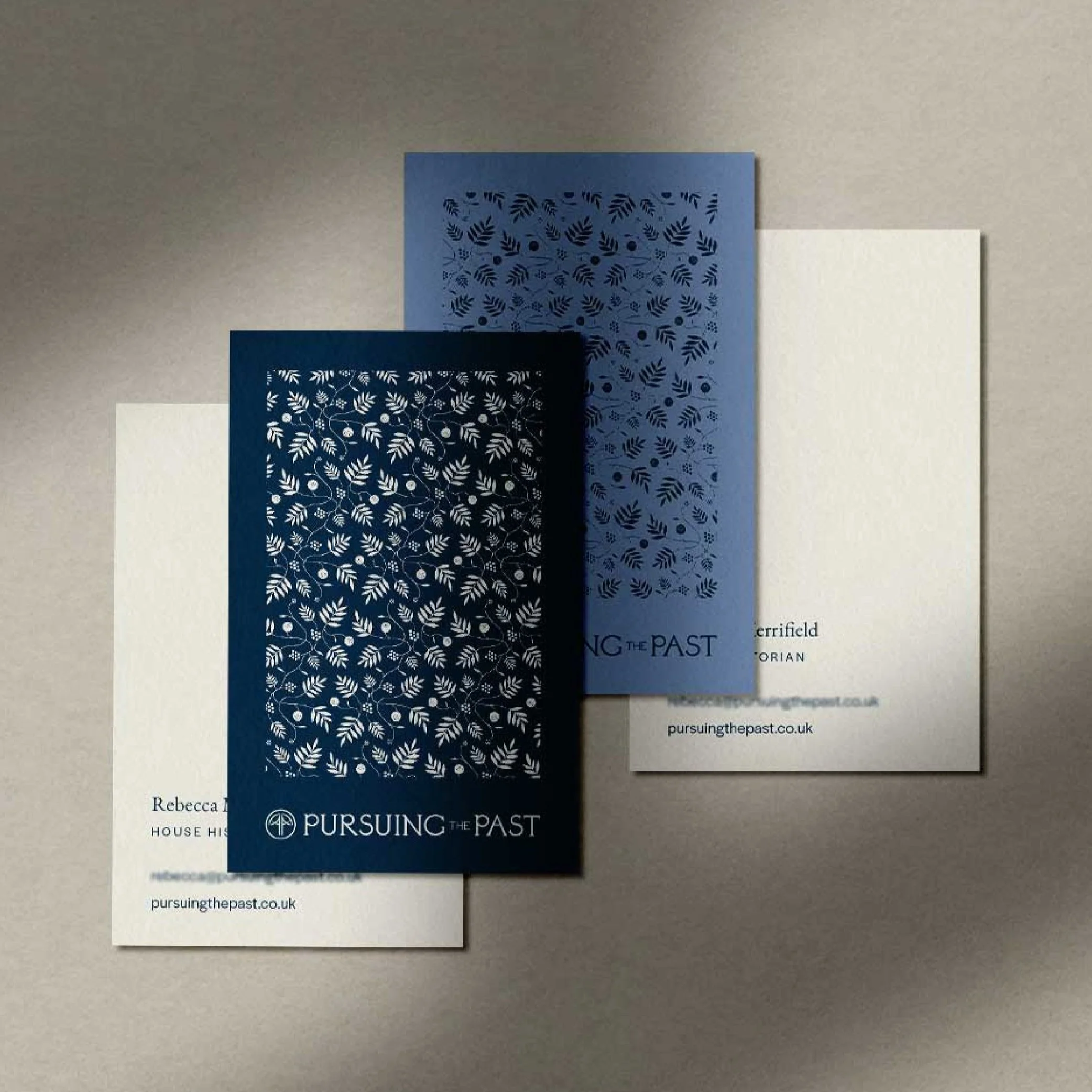 Business card design with floral designs, in shades of blue and white, arranged on a flat surface. The pamphlets are titled 'Pursuing the Past' and contain website information.