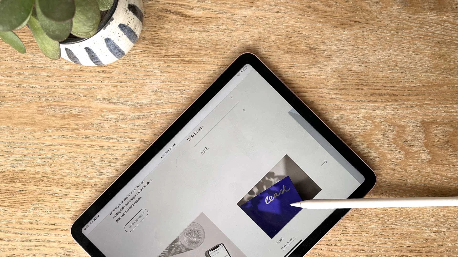 Tablet with stylus on wooden table next to a potted plant.