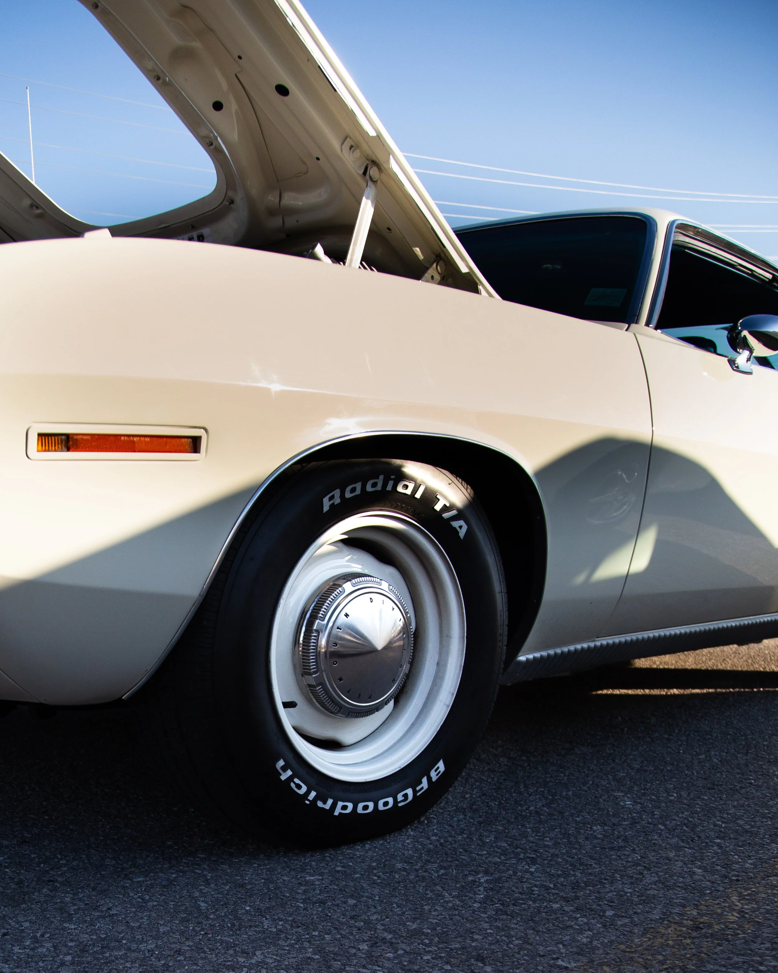 A close-up of a classic white vintage car with the hood open, showing a wheel with BFGoodrich Radial T/A tires, on a paved surface during daytime.