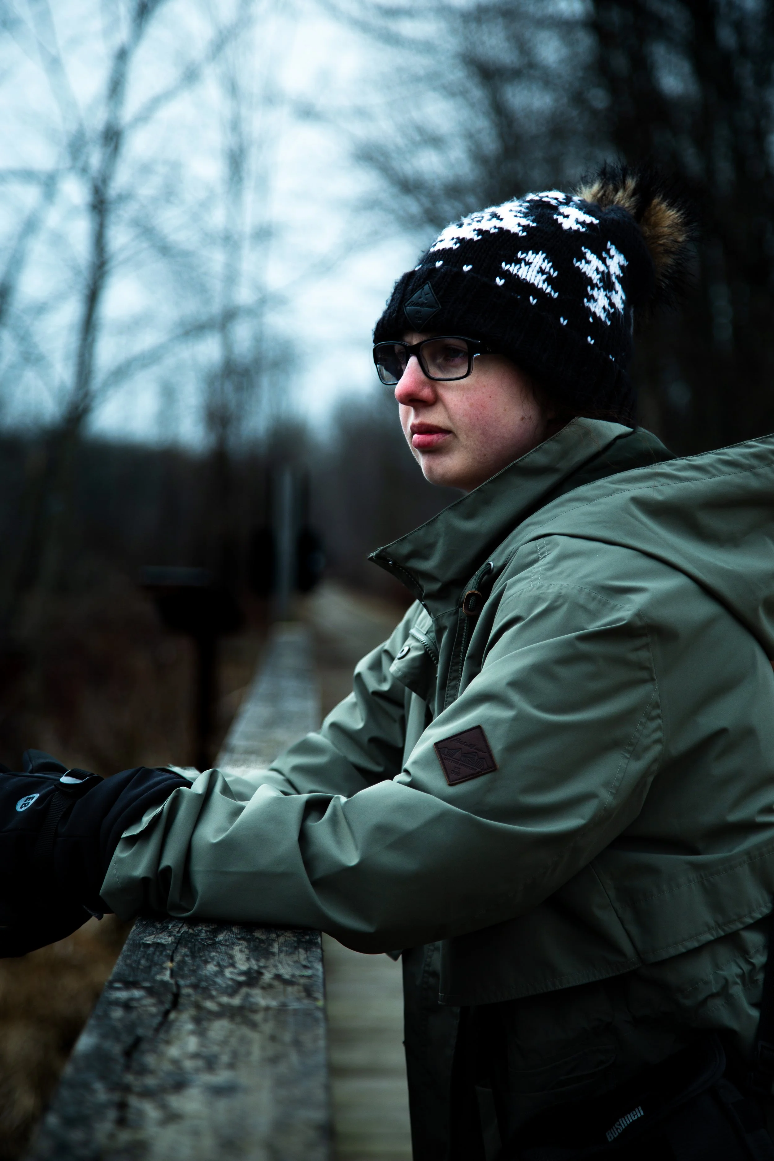 Person wearing glasses, a knit hat with snowflake pattern and pom-pom, and a green jacket, leaning on a wooden railing on a cloudy day in a wooded area.