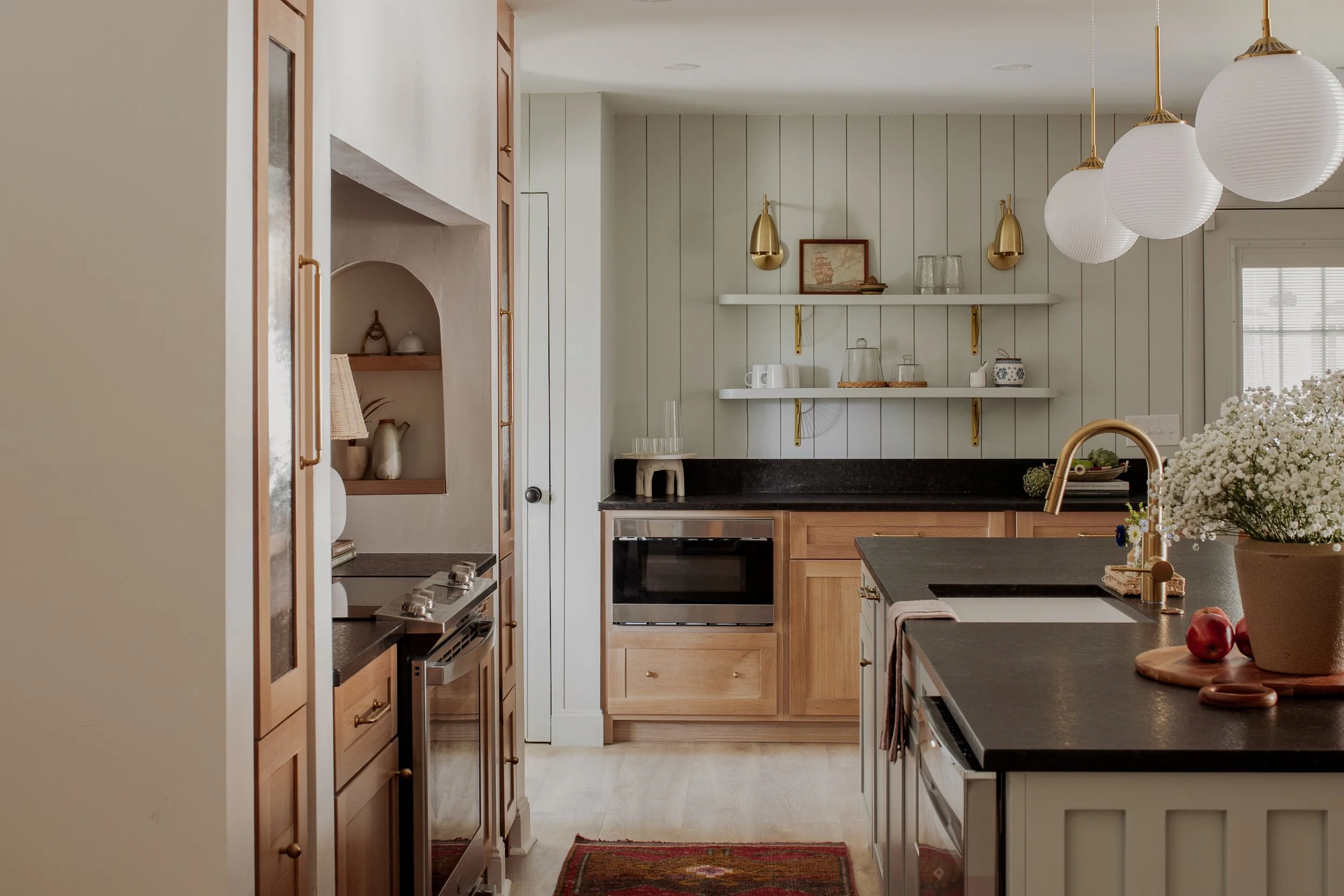 Modern kitchen with wooden cabinets, black countertops, and island with flowers. Brass fixtures and pendant lights, open shelves with decor and dishes.