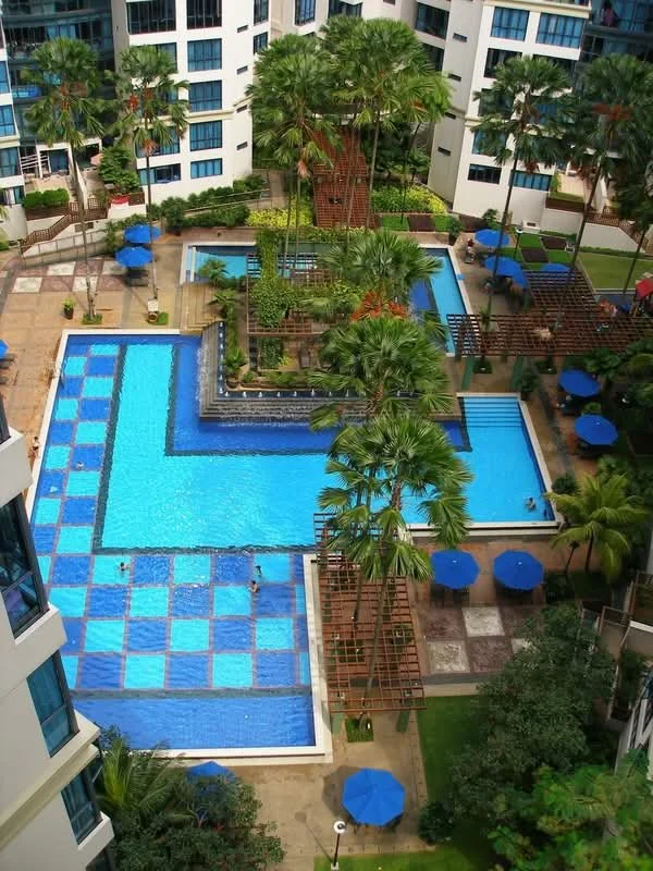Resort-style condominium swimming pool with blue-tiled sections, surrounded by palm trees, landscaped gardens, and mid-rise residential buildings.
