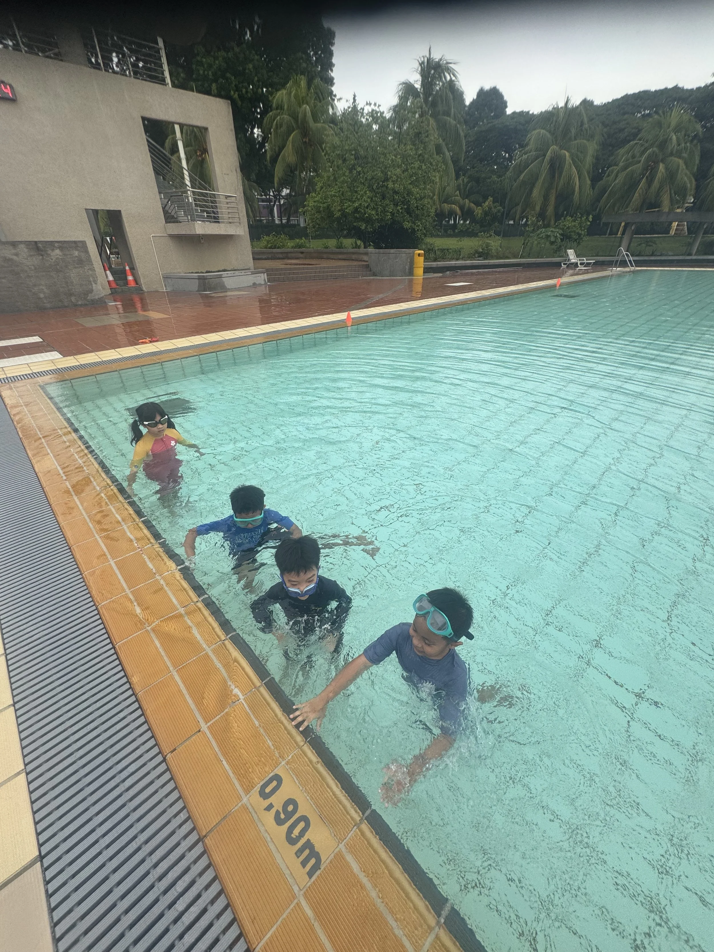 Children wearing flotation devices and masks swimming in an outdoor pool with a tiled deck, greenery, and trees in the background.