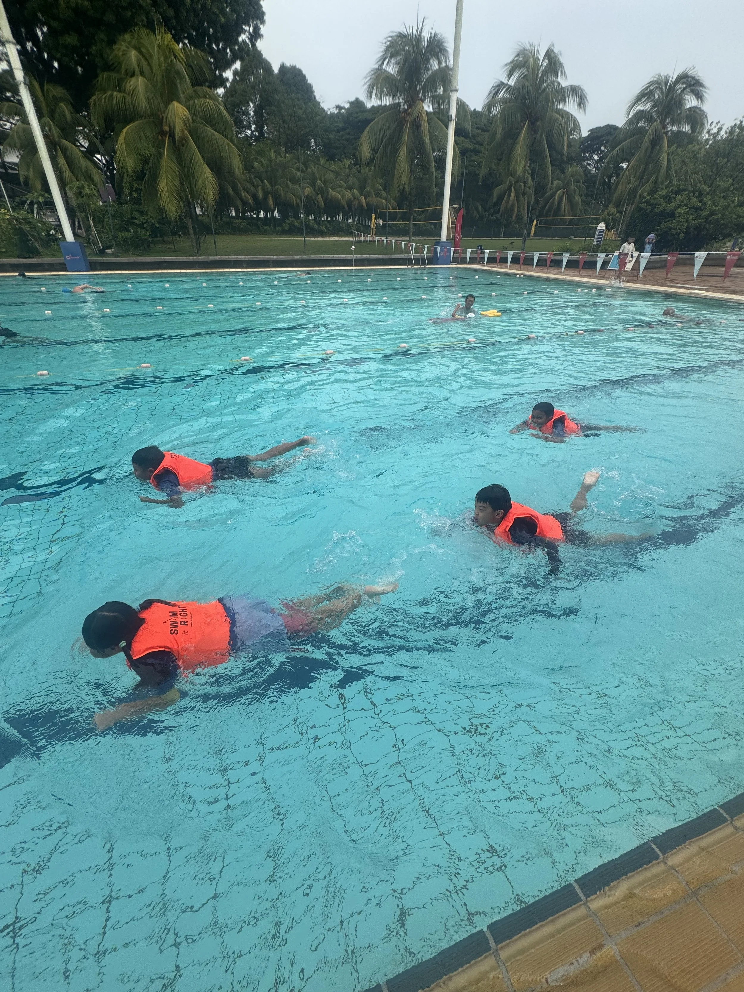 Children wearing orange life jackets swimming and practicing water safety in an outdoor pool surrounded by palm trees.