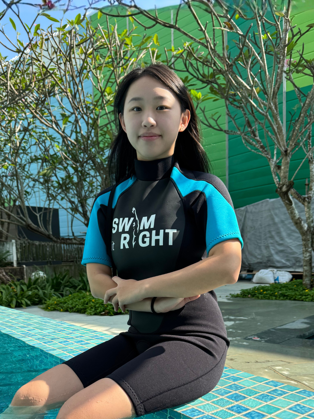 A young woman sitting on the edge of a swimming pool with her feet in the water, smiling, outdoors with trees and a building in the background.