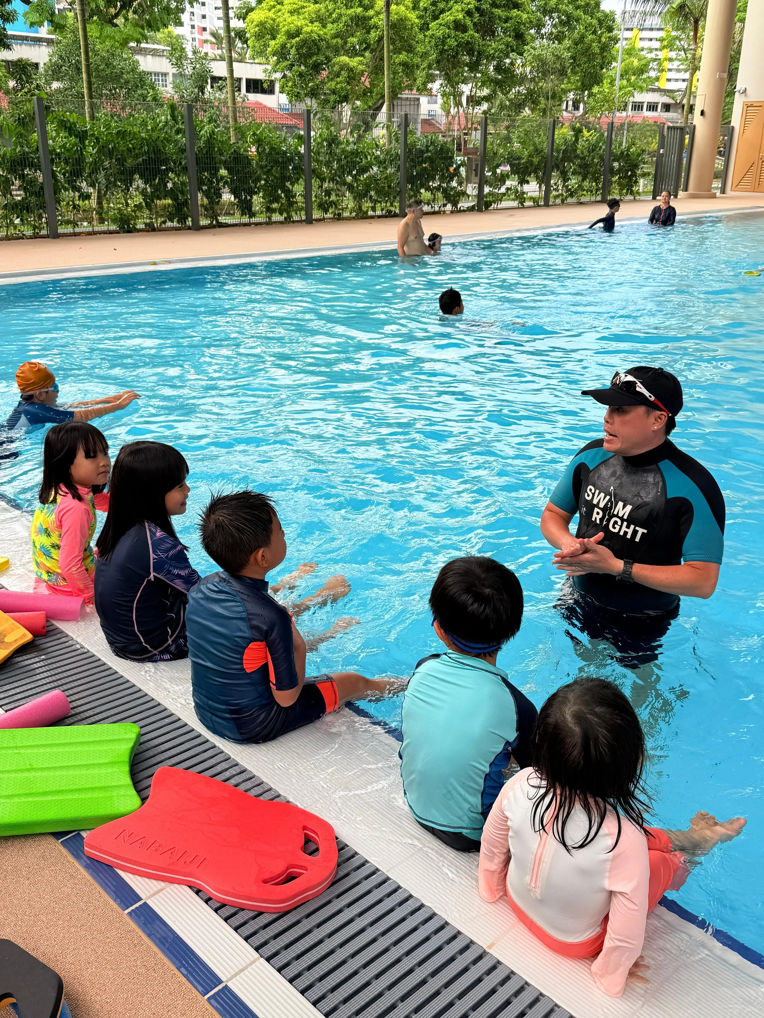 Children sitting by the edge of an indoor swimming pool while an instructor explains swimming techniques. Some children are in the water, and others are waiting on the poolside, with colorful flotation devices nearby, as part of a swimming lesson.