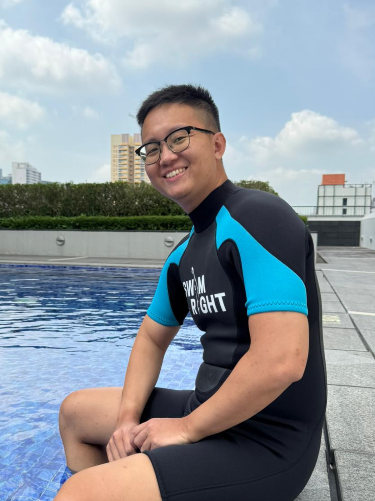A young man wearing glasses and a black and blue swimming wetsuit sits on the edge of a blue tiled pool, smiling at the camera with city buildings and a partly cloudy sky in the background.