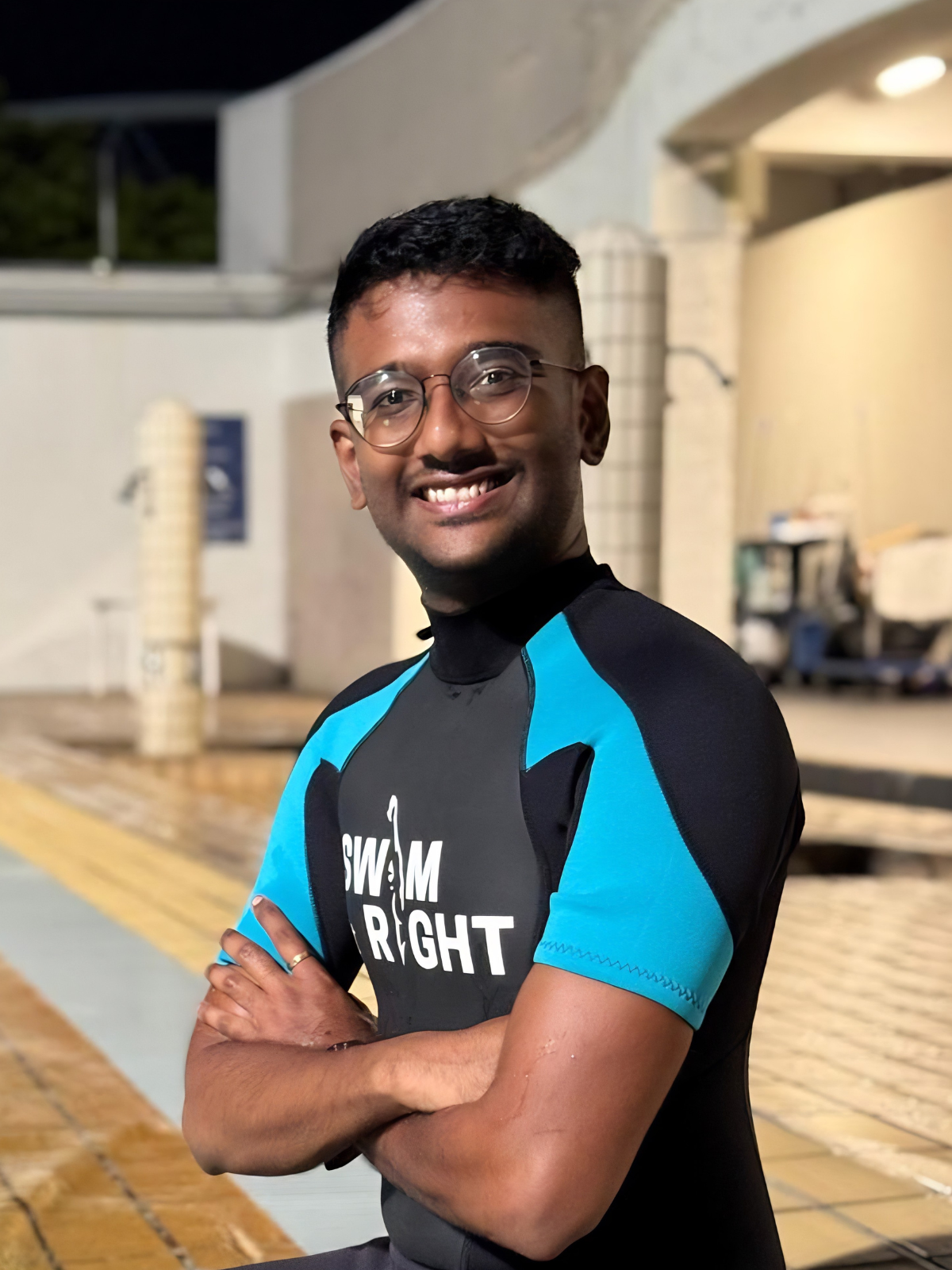 Young man sitting by the edge of a swimming pool at night, smiling, wearing a blue and black swim shirt with the text 'Swim Right,' and black swim shorts.